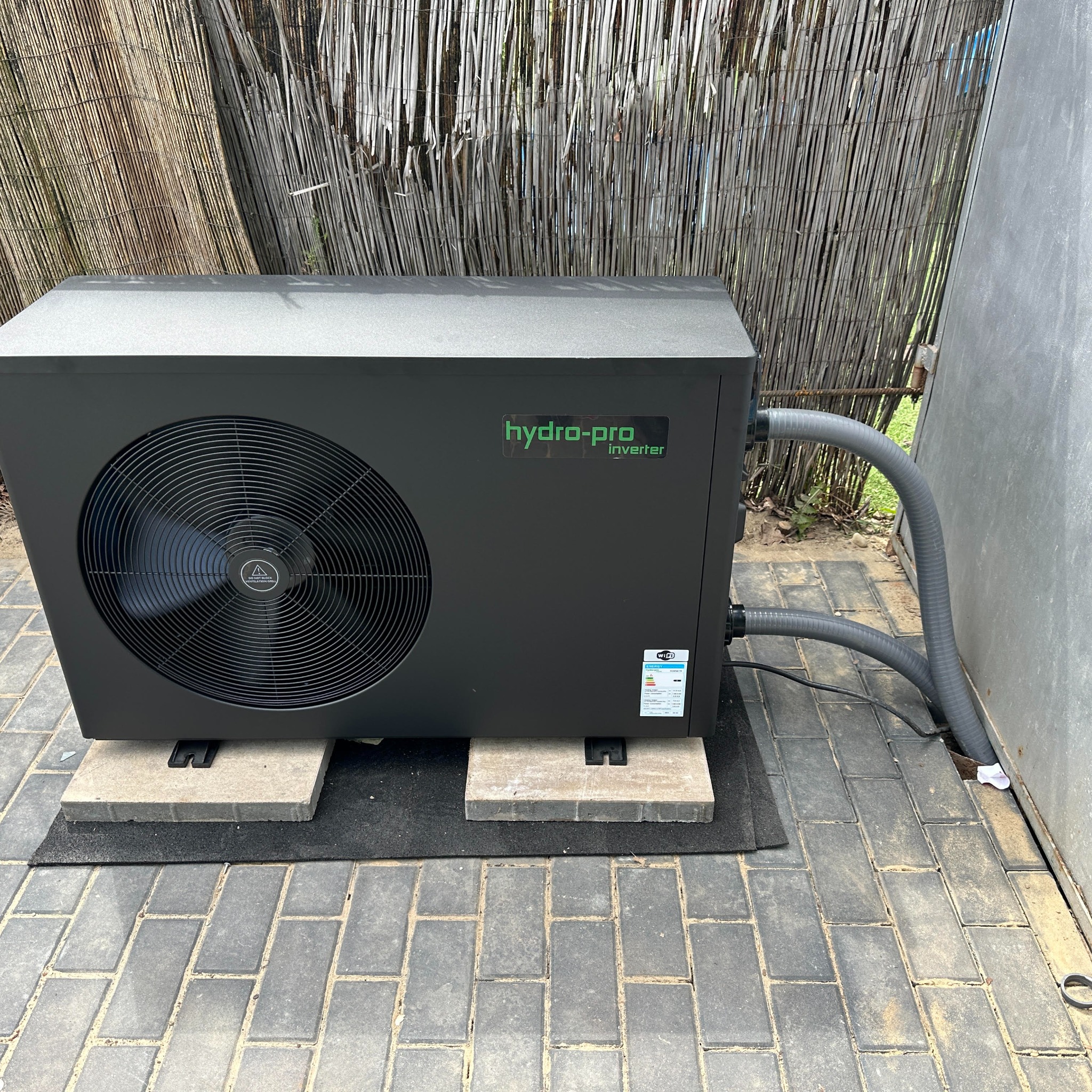 Czarna pompa ciepła Hydro-Pro Inverter, umieszczona na betonowych podstawach na kostce brukowej, z szarymi rurami podłączonymi z boku, na tle bambusowego ogrodzenia i szarej ściany.