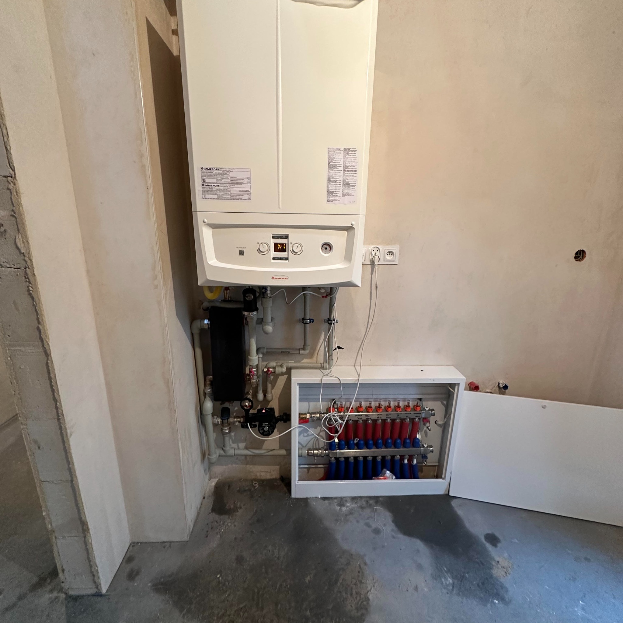 Biały piec gazowy marki Immergas zamontowany na ścianie w surowym pomieszczeniu, widoczna instalacja grzewcza i rozdzielacz z czerwonymi i niebieskimi rurami.