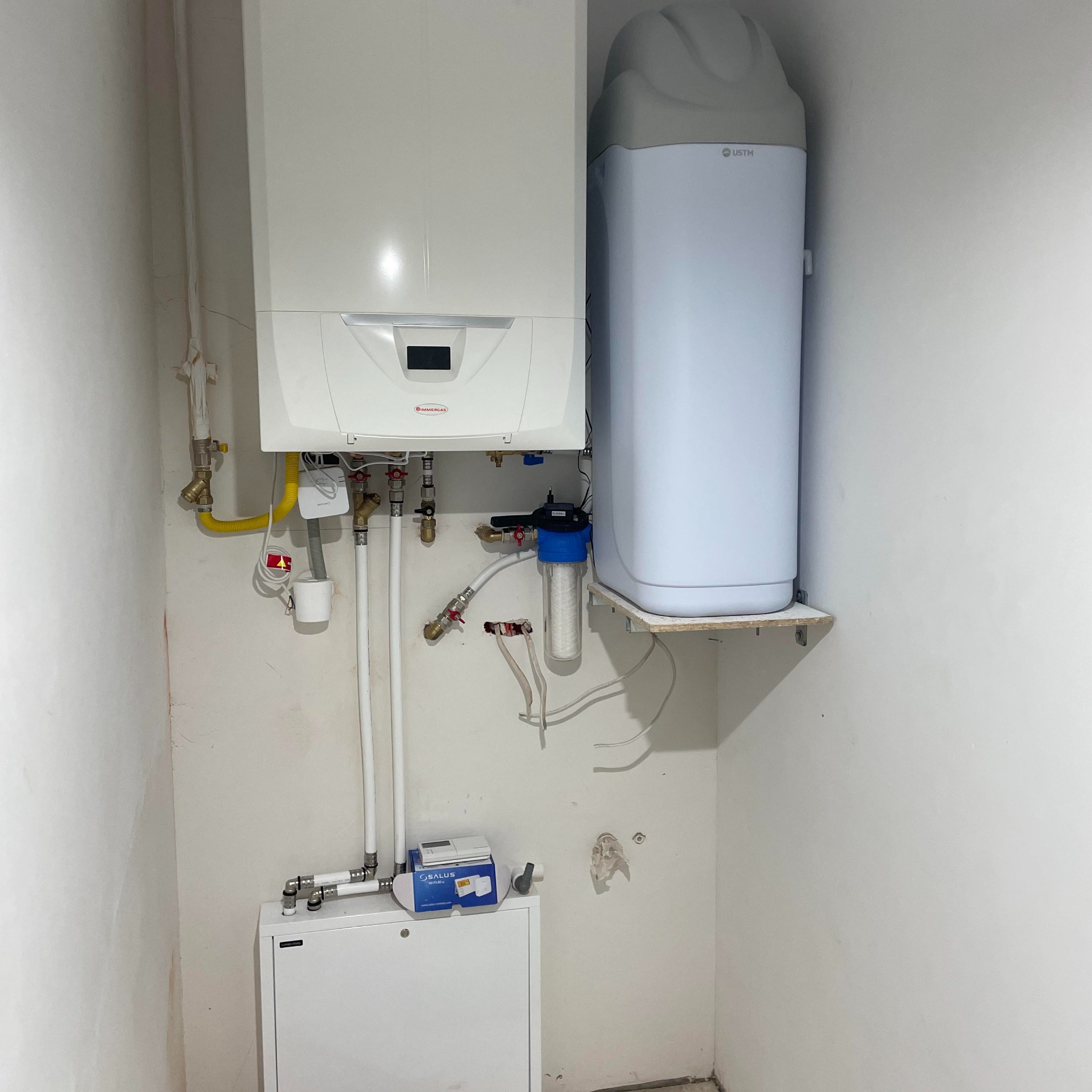 Biały piec gazowy marki Immergas z podłączonymi rurami i filtrem wody, obok zmiękczacz wody USTM na półce, widoczne elementy instalacji grzewczej i gazowej na tle białej ściany.
