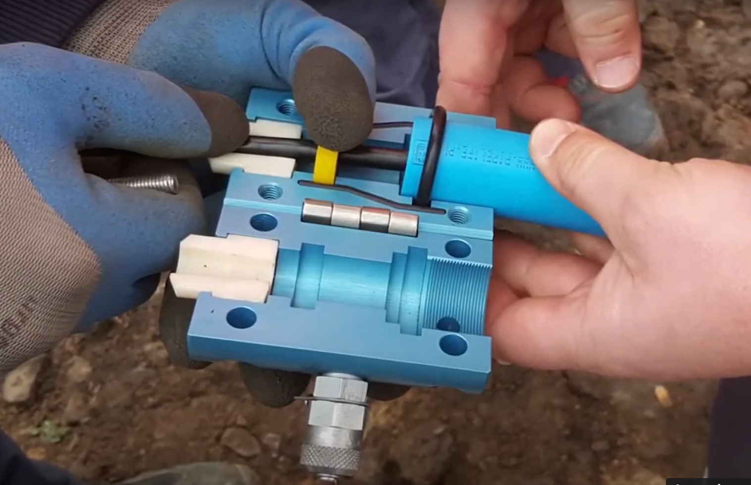 Instalator w rękawicach łączy niebieską rurę z metalowym łącznikiem, widoczne przewody i elementy mocujące.