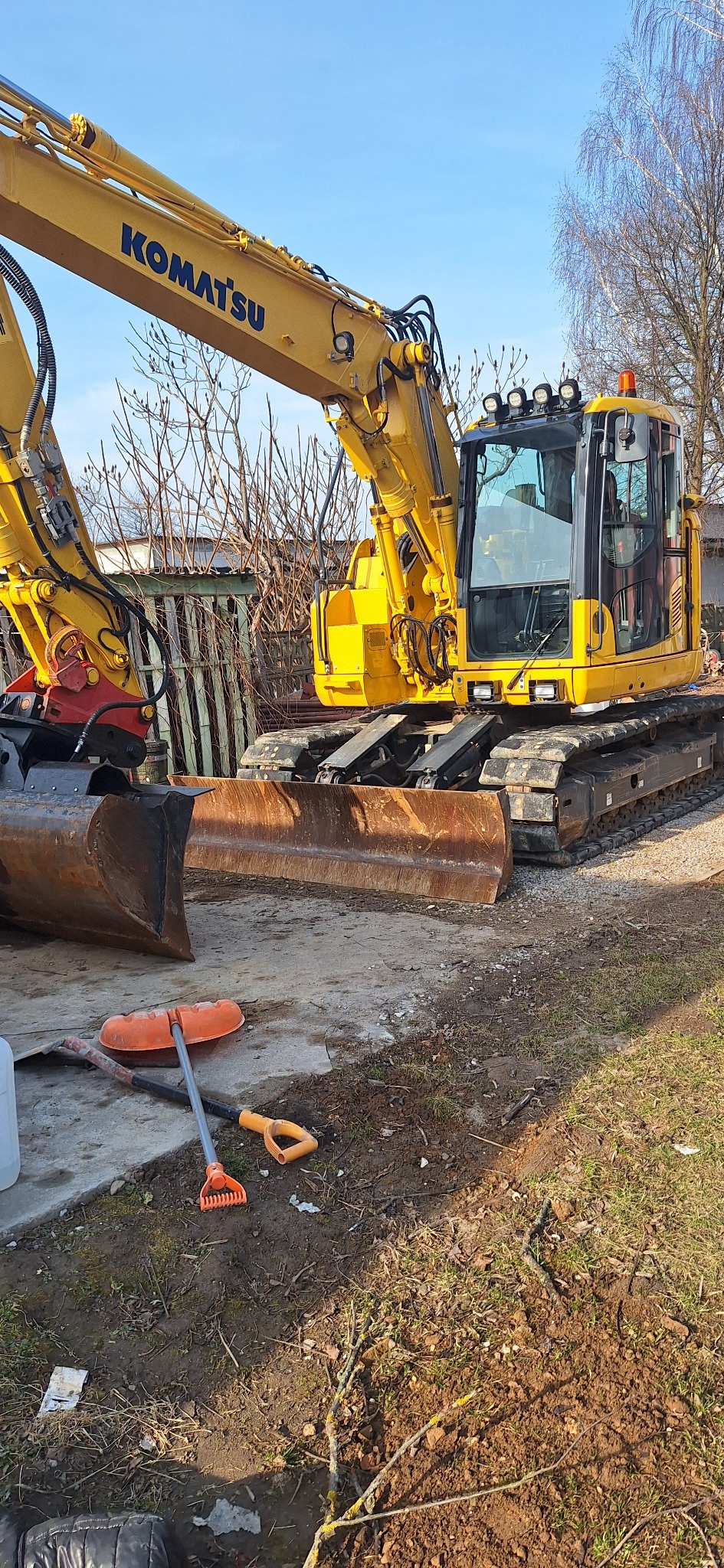 Żółta koparka Komatsu na placu budowy, w tle drzewa bez liści i ogrodzenie, obok leżą łopaty i inne narzędzia.