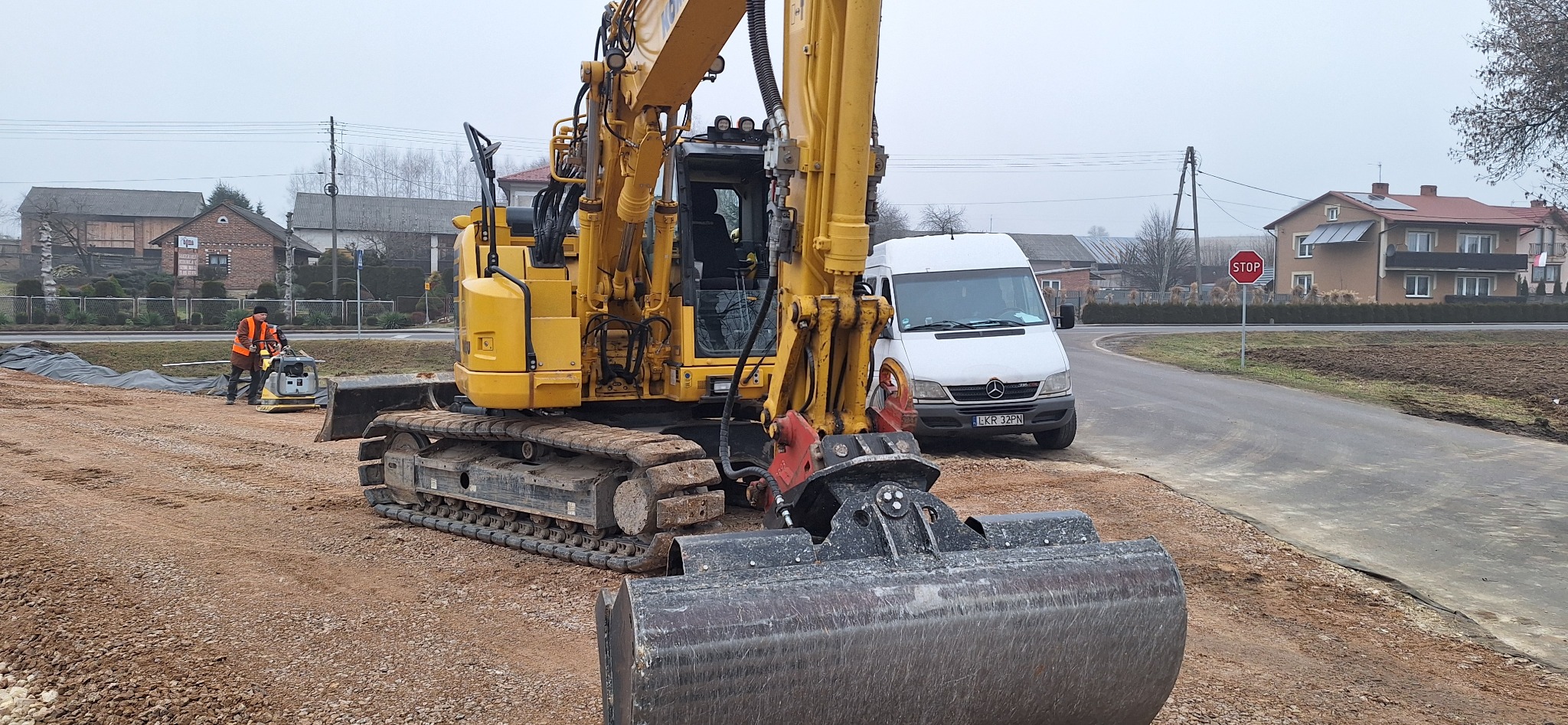 Żółta koparka i ubijarka gruntu na placu budowy; w tle biały bus i znak STOP