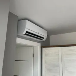 Biały klimatyzator Mitsubishi zamontowany na ścianie nad drzwiami i szafą z żaluzjowymi drzwiami. Szara ściana. Wnętrze mieszkania.