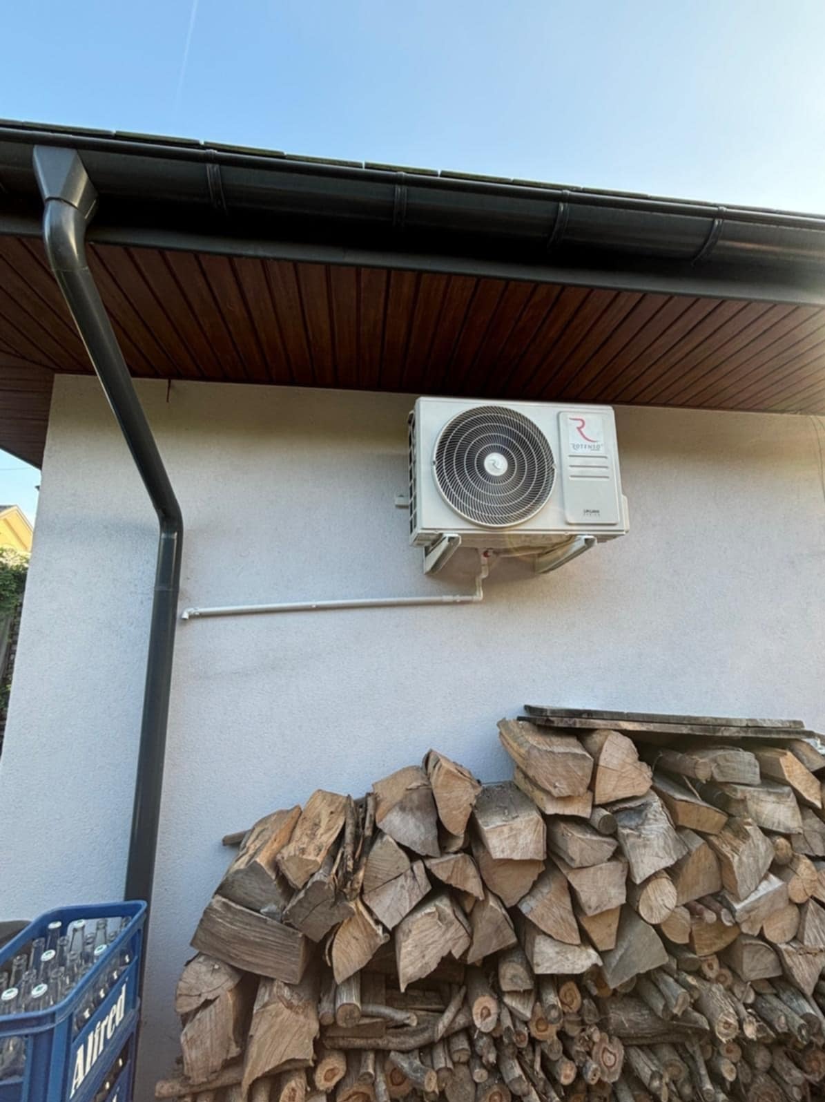 Zamontowany klimatyzator Rotenso na elewacji domu, z widoczną stertą drewna i rynną. Ujęcie w słoneczny dzień.