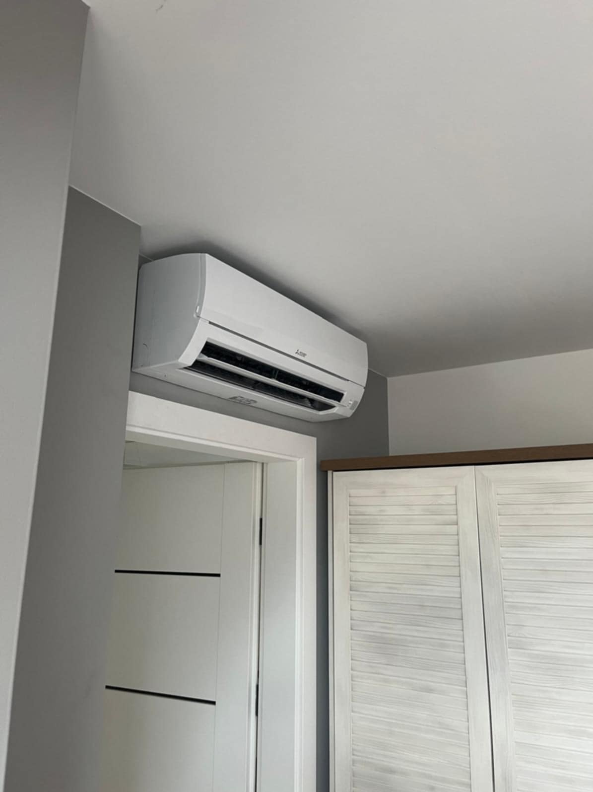 Biały klimatyzator Mitsubishi zamontowany na ścianie nad drzwiami i szafą z żaluzjowymi drzwiami. Szara ściana. Wnętrze mieszkania.