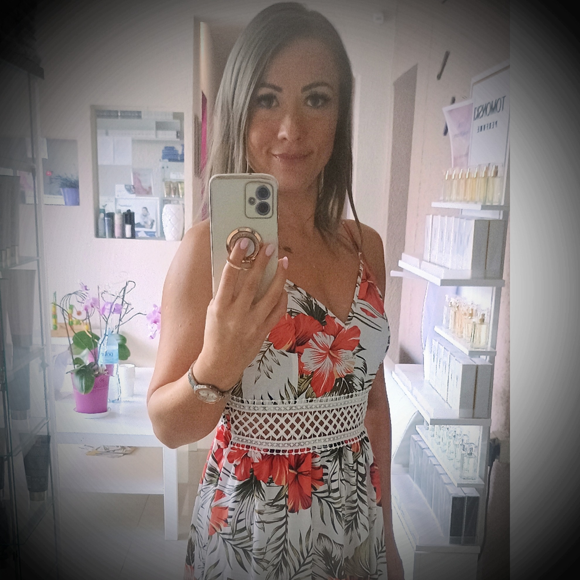 Kobieta robi selfie w salonie kosmetycznym, w tle półki z perfumami i kwiaty.