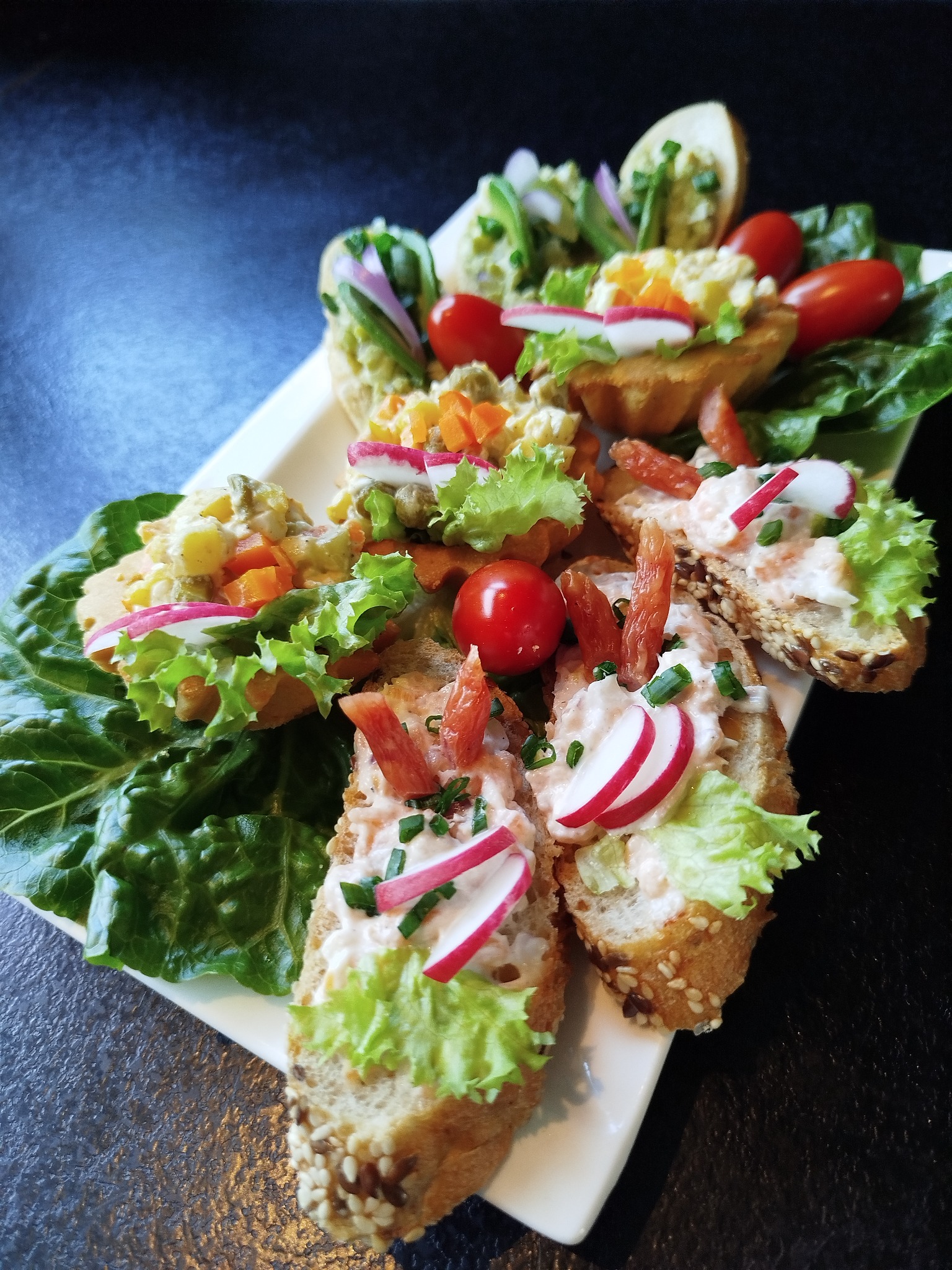 Elegancko podane kanapki na białej tacy, udekorowane sałatą, rzodkiewką i pomidorami koktajlowymi, z różnymi pastami i dodatkami, idealne na przyjęcie.