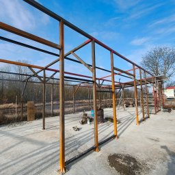 PRO-GRUPA SPÓŁKA Z OGRANICZONĄ ODPOWIEDZIALNOŚCIĄ - Konstrukcja stalowa w trakcie budowy, pomalowana na pomarańczowo i czarno, na tle błękitnego nieba. Widoczne narzędzia i elementy placu budowy.