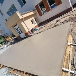 PRO-GRUPA SPÓŁKA Z OGRANICZONĄ ODPOWIEDZIALNOŚCIĄ - Świeżo wylana, gładka wylewka betonowa na placu budowy, otoczona drewnianym szalunkiem. W tle budynek z oknami i pracownik budowlany. Widoczne narzędzia i materiały budowlane.