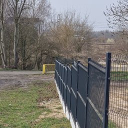 PRO-GRUPA SPÓŁKA Z OGRANICZONĄ ODPOWIEDZIALNOŚCIĄ - Nowoczesne ogrodzenie panelowe w kolorze grafitowym, zamontowane na betonowej podmurówce. Widok z boku, z perspektywą zbiegającą się w oddali. Trawa i drzewa w tle.