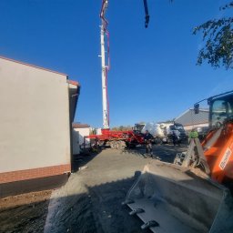PRO-GRUPA SPÓŁKA Z OGRANICZONĄ ODPOWIEDZIALNOŚCIĄ - Betoniarka pompuje beton na budowę domu jednorodzinnego. Widoczny operator, koparka i fragment budynku z ceglaną podmurówką. Błękitne niebo w tle.