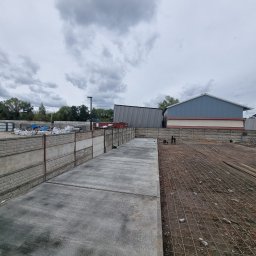 PRO-GRUPA SPÓŁKA Z OGRANICZONĄ ODPOWIEDZIALNOŚCIĄ - Betonowa wylewka na placu budowy, widoczne zbrojenia i pracownicy w tle. Szare niebo i budynki w tle. Betonowa ściana oddzielająca teren.