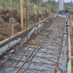 PRO-GRUPA SPÓŁKA Z OGRANICZONĄ ODPOWIEDZIALNOŚCIĄ - Zbrojenie fundamentu w wykopie z deskowaniem i folią hydroizolacyjną. Widoczne pręty zbrojeniowe i drewniane elementy szalunku. Prace ziemne w tle.