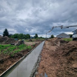 PRO-GRUPA SPÓŁKA Z OGRANICZONĄ ODPOWIEDZIALNOŚCIĄ - Wylewka fundamentowa w szalunku, beton rozprowadzany pompą. Widoczne zbrojenie i przygotowany teren budowy pod pochmurnym niebem.