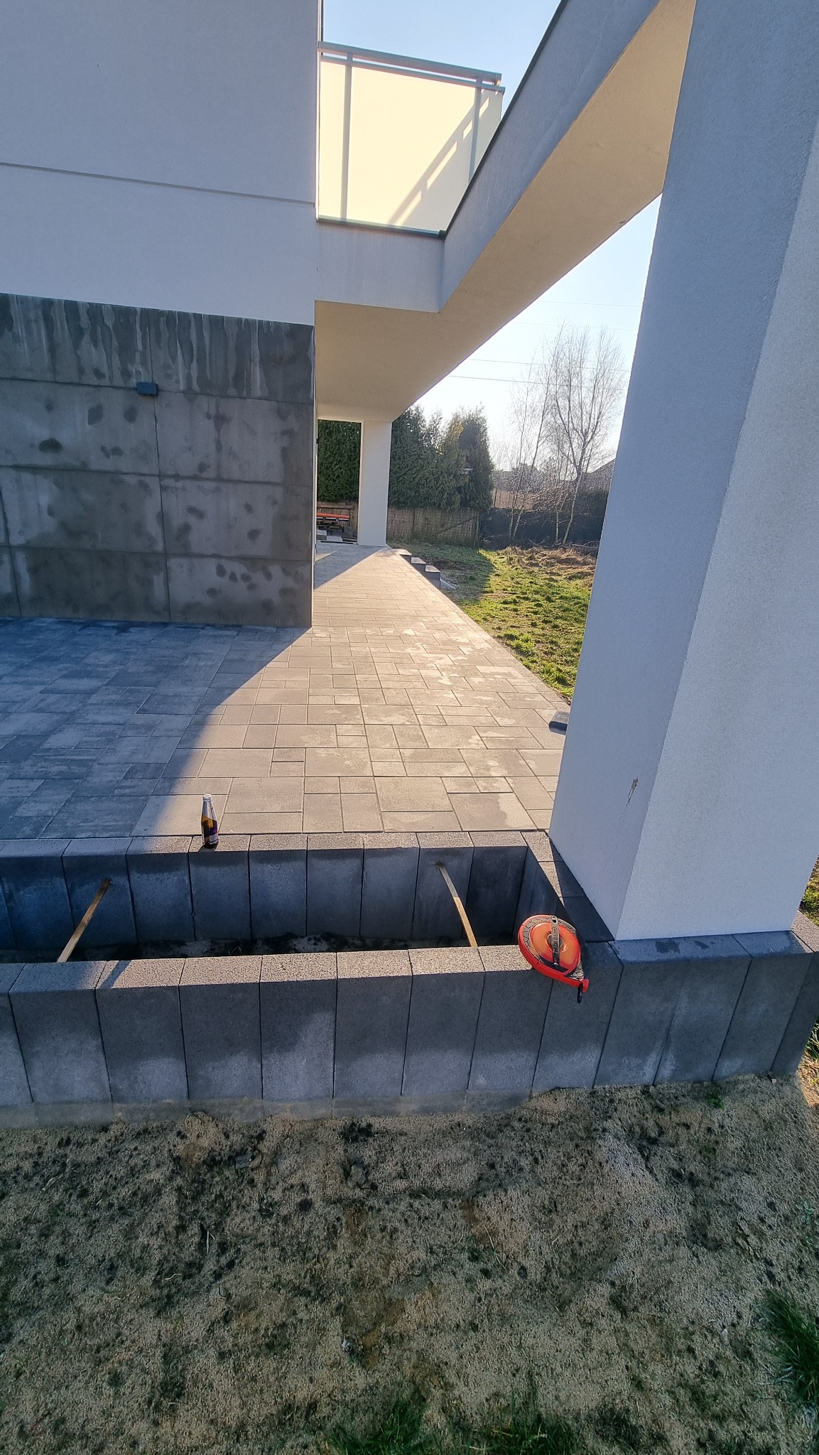 Nowoczesny taras z szarej kostki brukowej w trakcie budowy, widoczne podpory i poziomica, tło stanowi budynek z białymi elementami architektonicznymi.