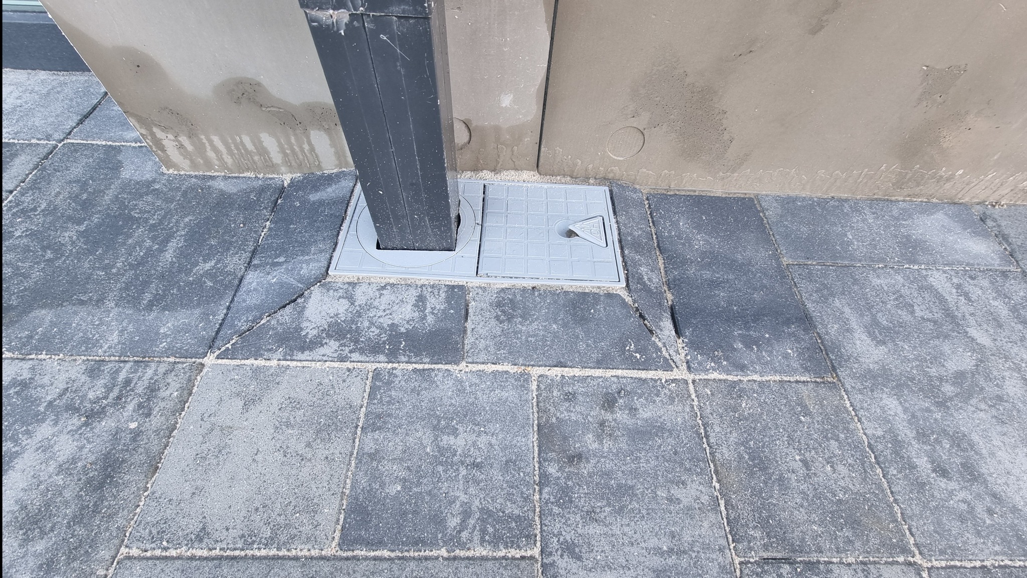 Szara kostka brukowa ułożona wokół słupka z zamontowaną szarą pokrywą studzienki rewizyjnej z fakturą kratki i trójkątnym uchwytem, widoczne ślady zaprawy na sąsiadującej ścianie.