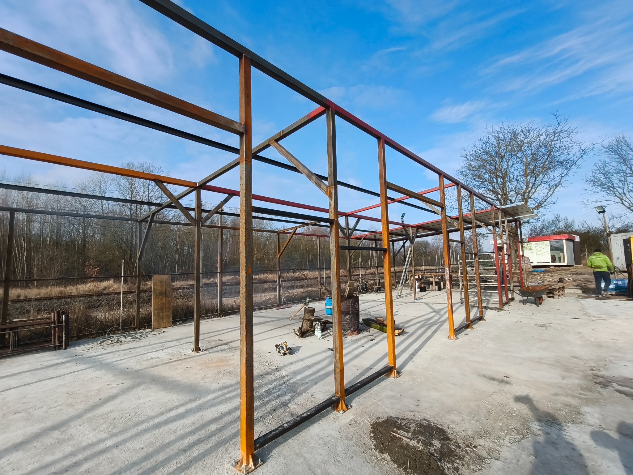 Konstrukcja stalowa w trakcie budowy, pomalowana na pomarańczowo i czarno, na tle błękitnego nieba. Widoczne narzędzia i elementy placu budowy.