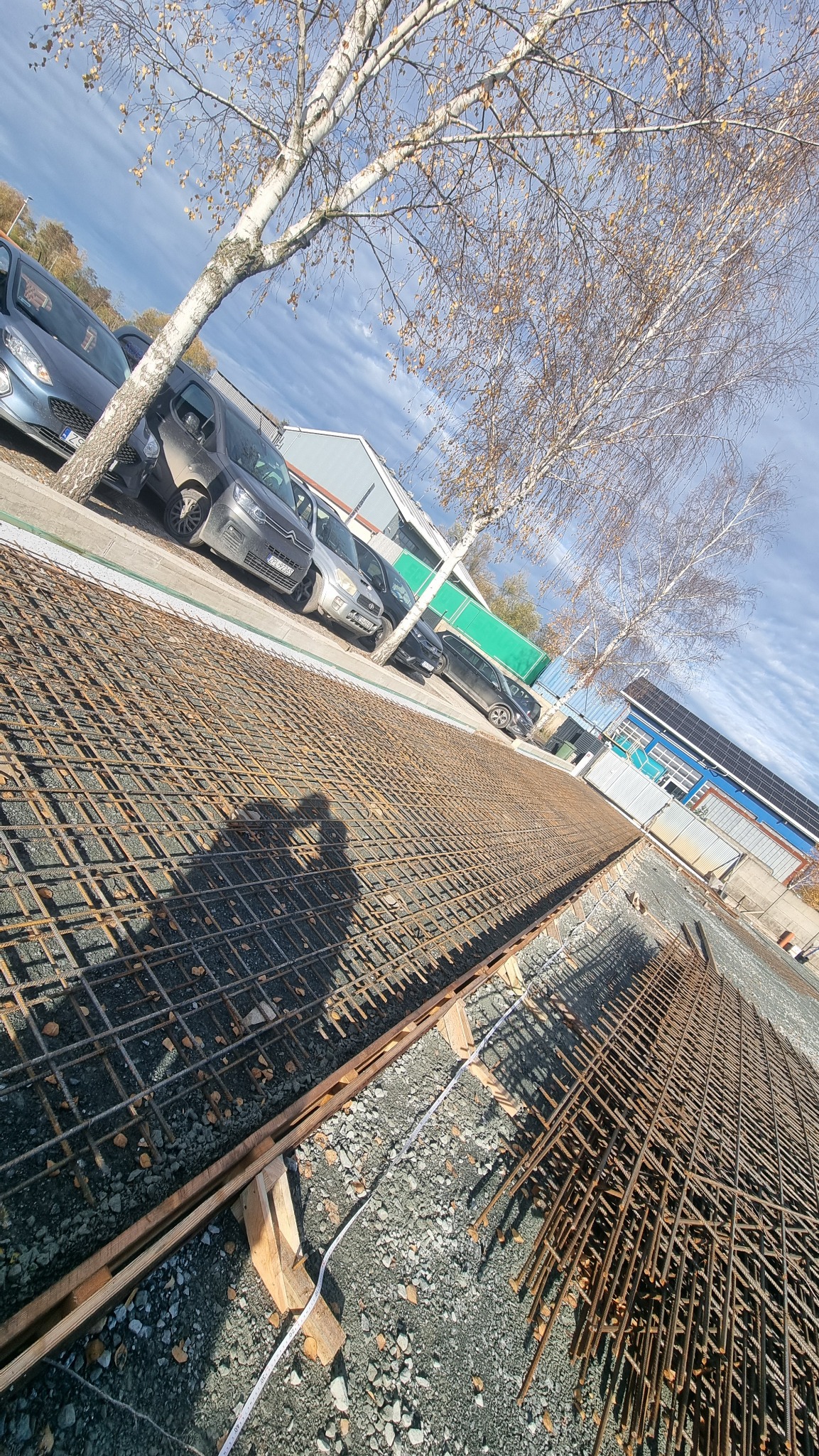 Siatka zbrojeniowa ułożona na żwirze, przygotowana do zalania betonem. W tle zaparkowane samochody i budynek z panelami słonecznymi. Widoczny cień dwóch osób.