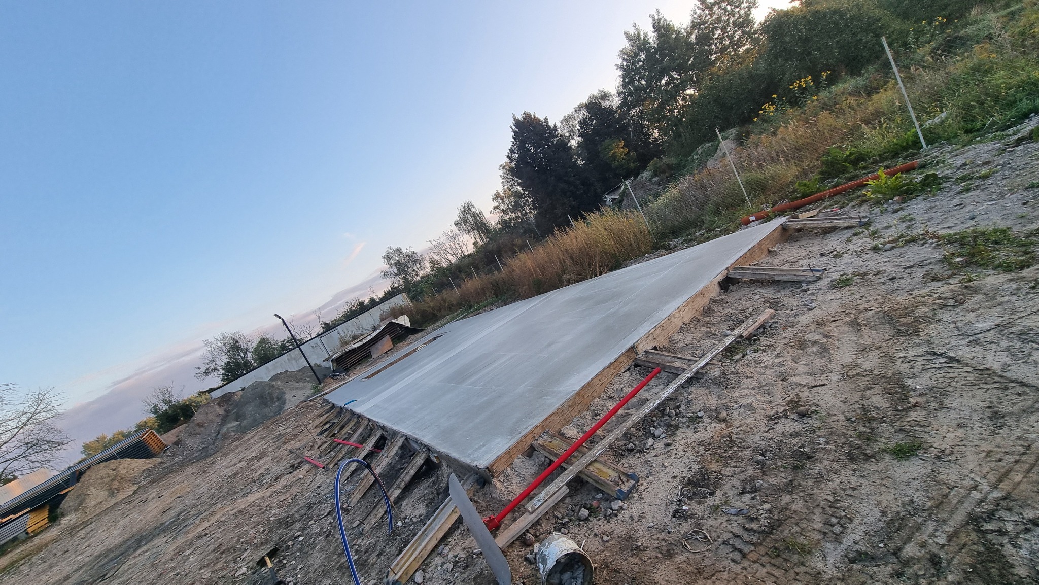 Świeżo wylana, gładka wylewka fundamentowa na nierównym terenie, otoczona drewnianym szalunkiem i narzędziami budowlanymi. W tle zieleń i błękitne niebo.