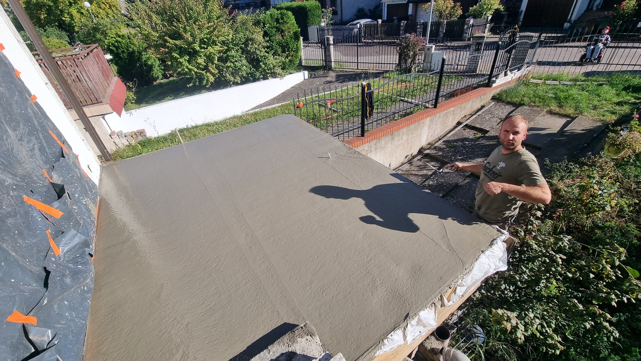 Świeżo wylana, gładka wylewka na zewnątrz budynku, z widocznym pracownikiem trzymającym kielnię i elementami otoczenia, takimi jak roślinność i ogrodzenie.