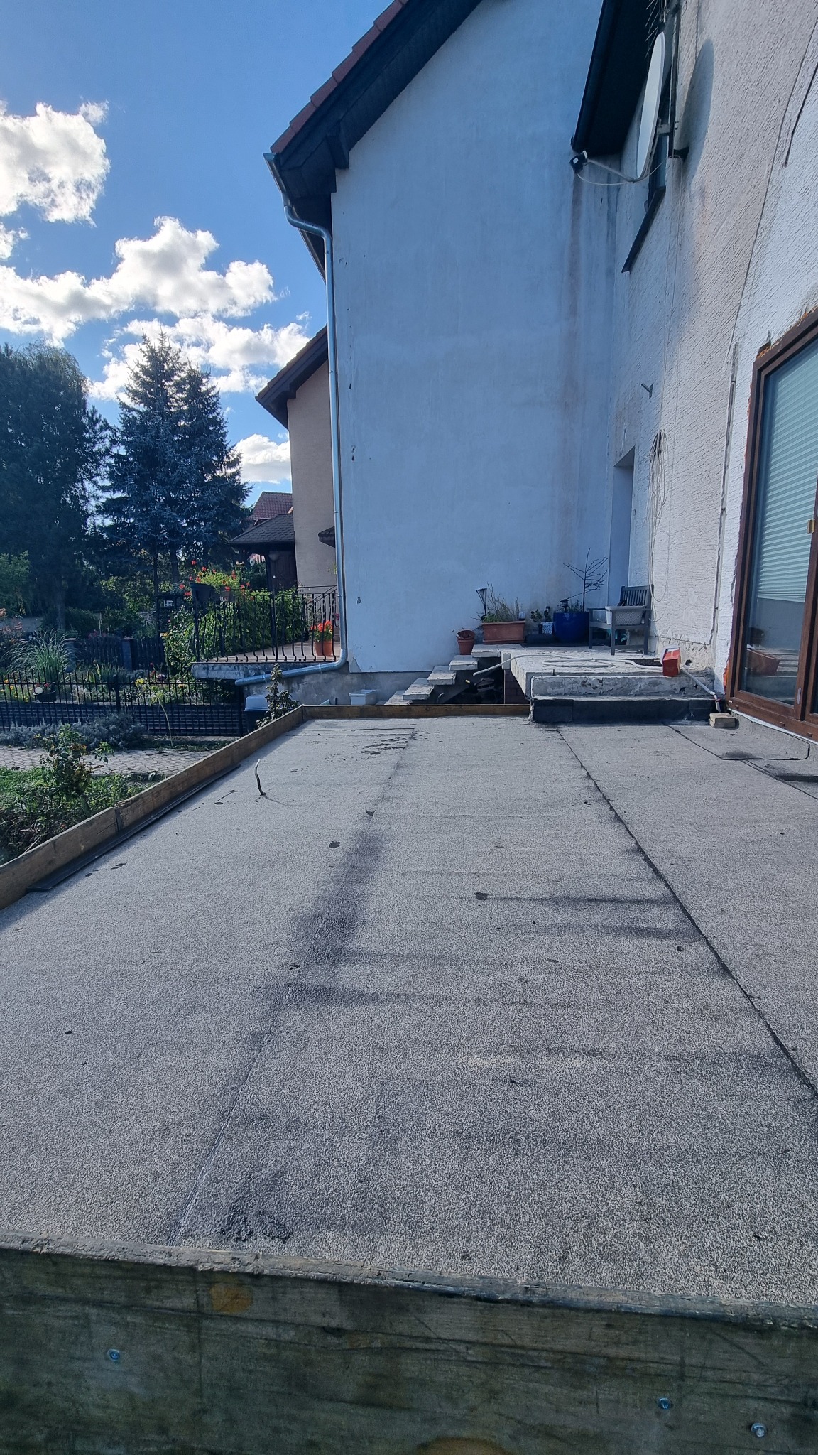 Wykończony, płaski dach budynku z widoczną papą i drewnianą obudową. W tle fragment ogrodu i elewacja budynku z oknem. Słoneczny dzień.