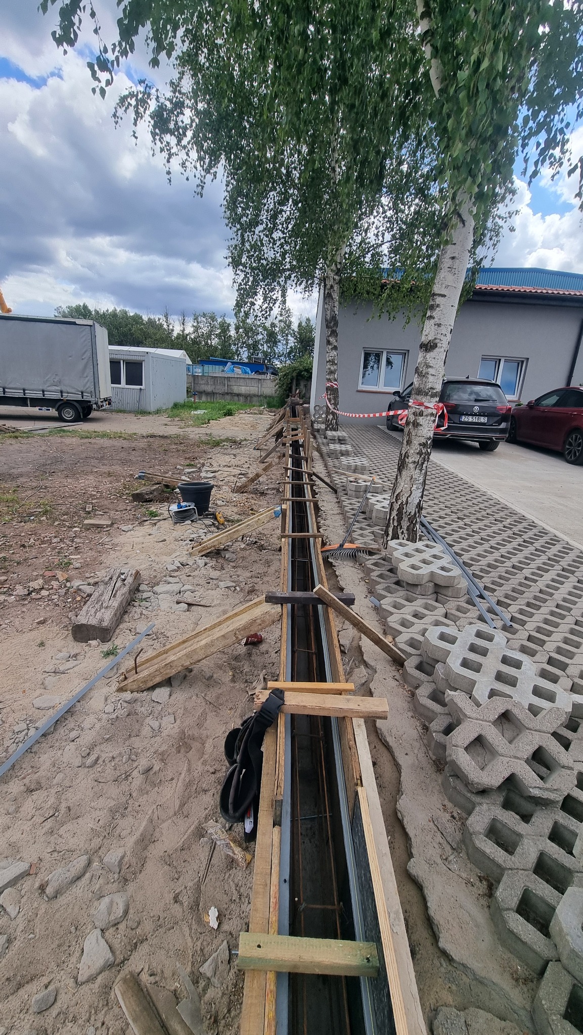 Przygotowanie szalunku i zbrojenia pod fundament liniowy, widoczne drewniane elementy konstrukcyjne i stalowe pręty. W tle budynek i drzewa.