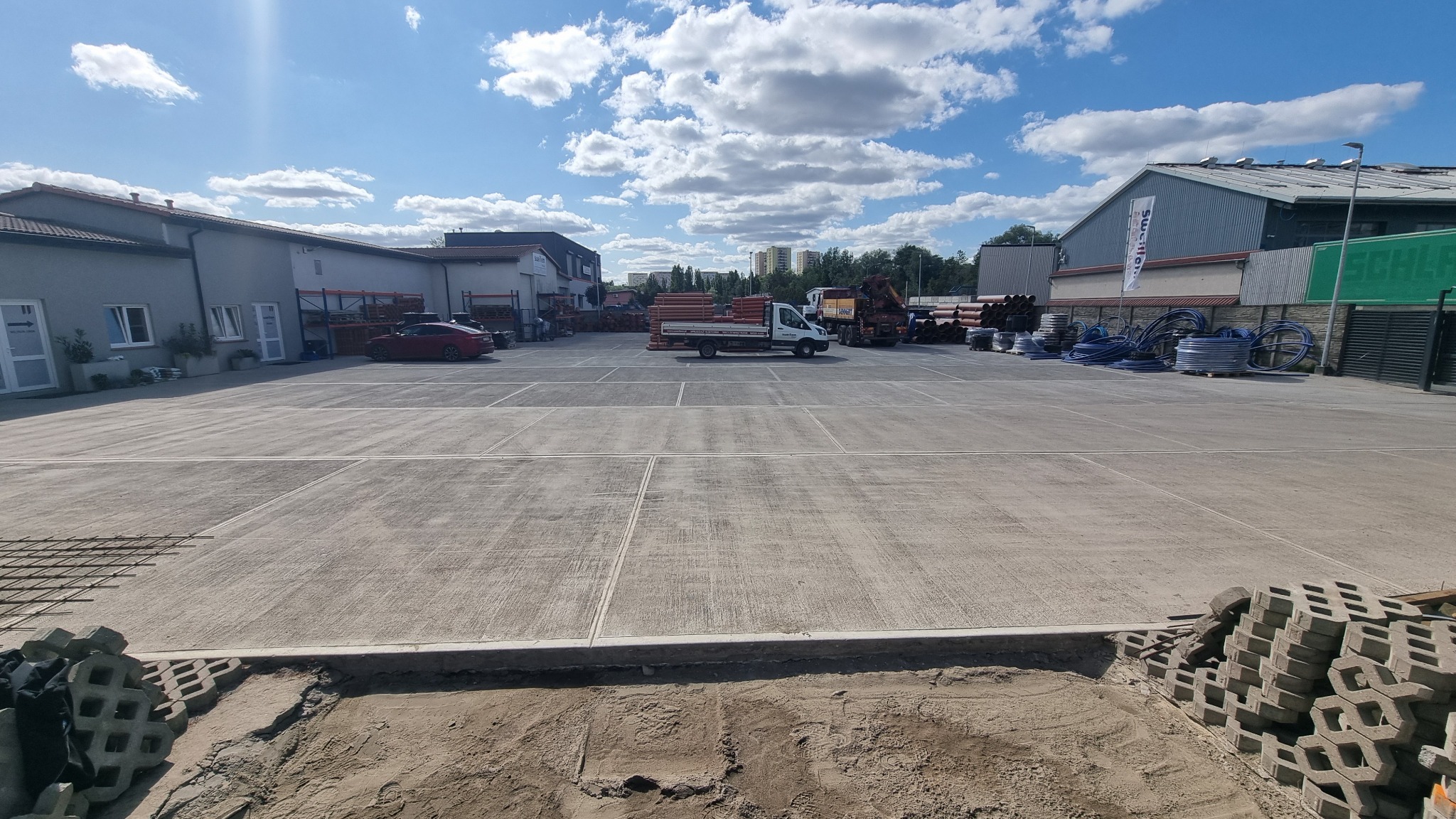 Rozległy plac budowy z betonową nawierzchnią, przygotowany pod wylewkę. Widoczne materiały budowlane, ciężarówka i budynki w tle, sugerujące etap prac ziemnych i przygotowawczych.