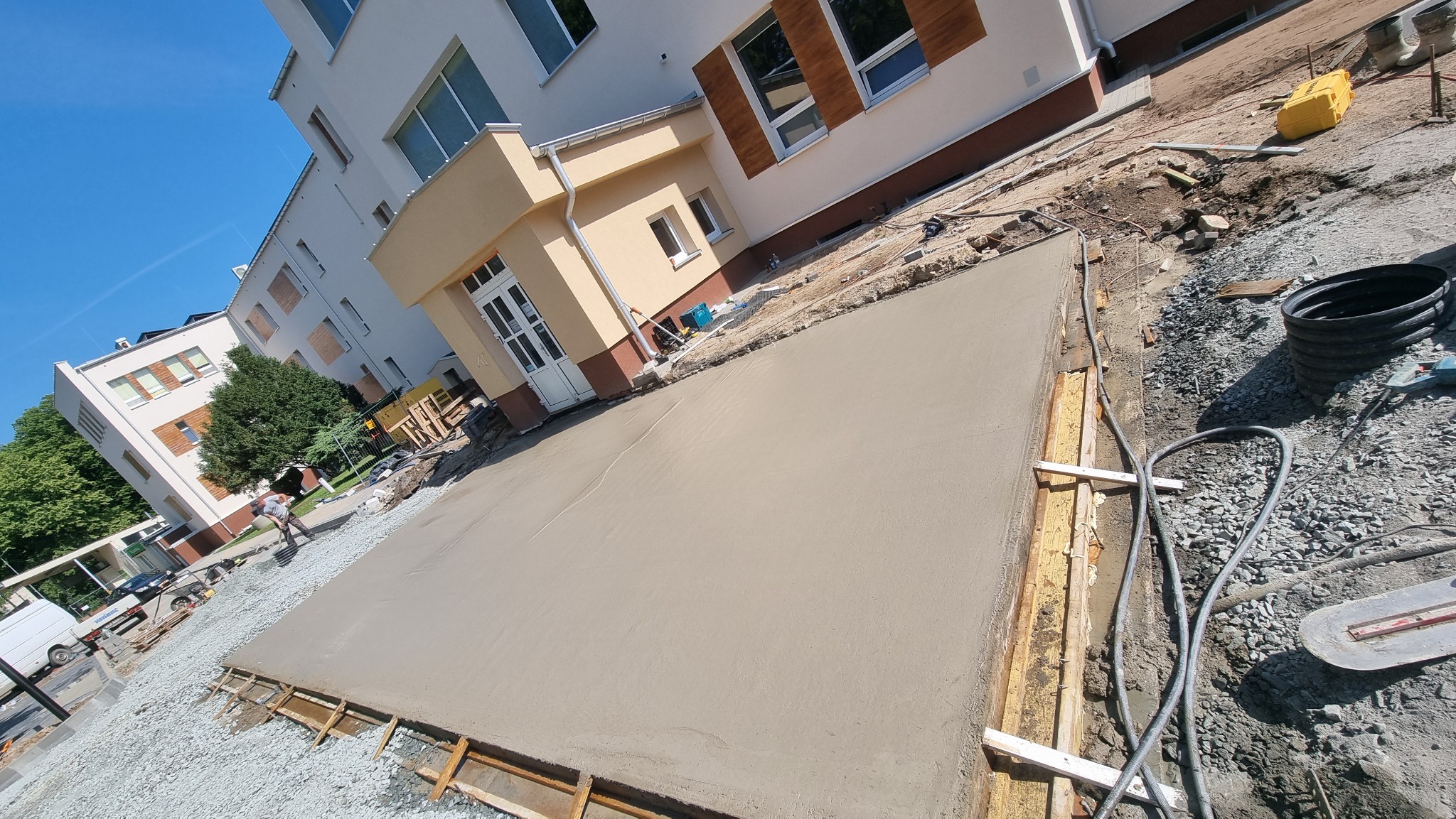 Świeżo wylana, gładka wylewka betonowa na placu budowy, otoczona drewnianym szalunkiem. W tle budynek z oknami i pracownik budowlany. Widoczne narzędzia i materiały budowlane.