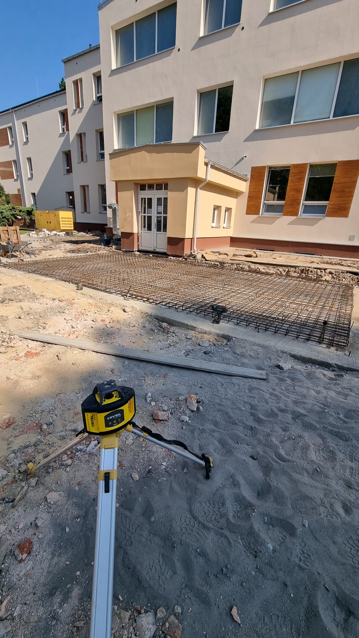 Przygotowanie terenu pod wylewkę: siatka zbrojeniowa rozłożona przed budynkiem, niwelator laserowy na statywie. Słoneczny dzień.