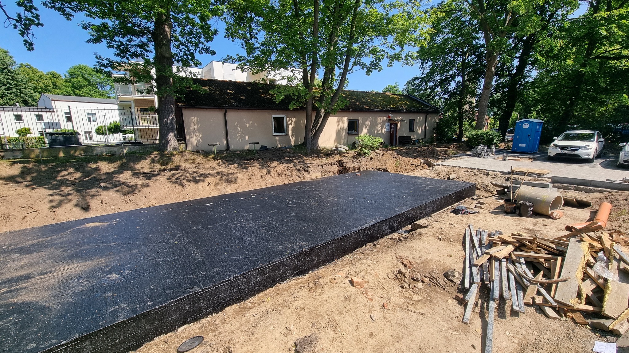 Zabezpieczony fundament budynku w wykopie, widoczne materiały budowlane i fragment budynku w tle. Ujęcie z poziomu gruntu, w słoneczny dzień.