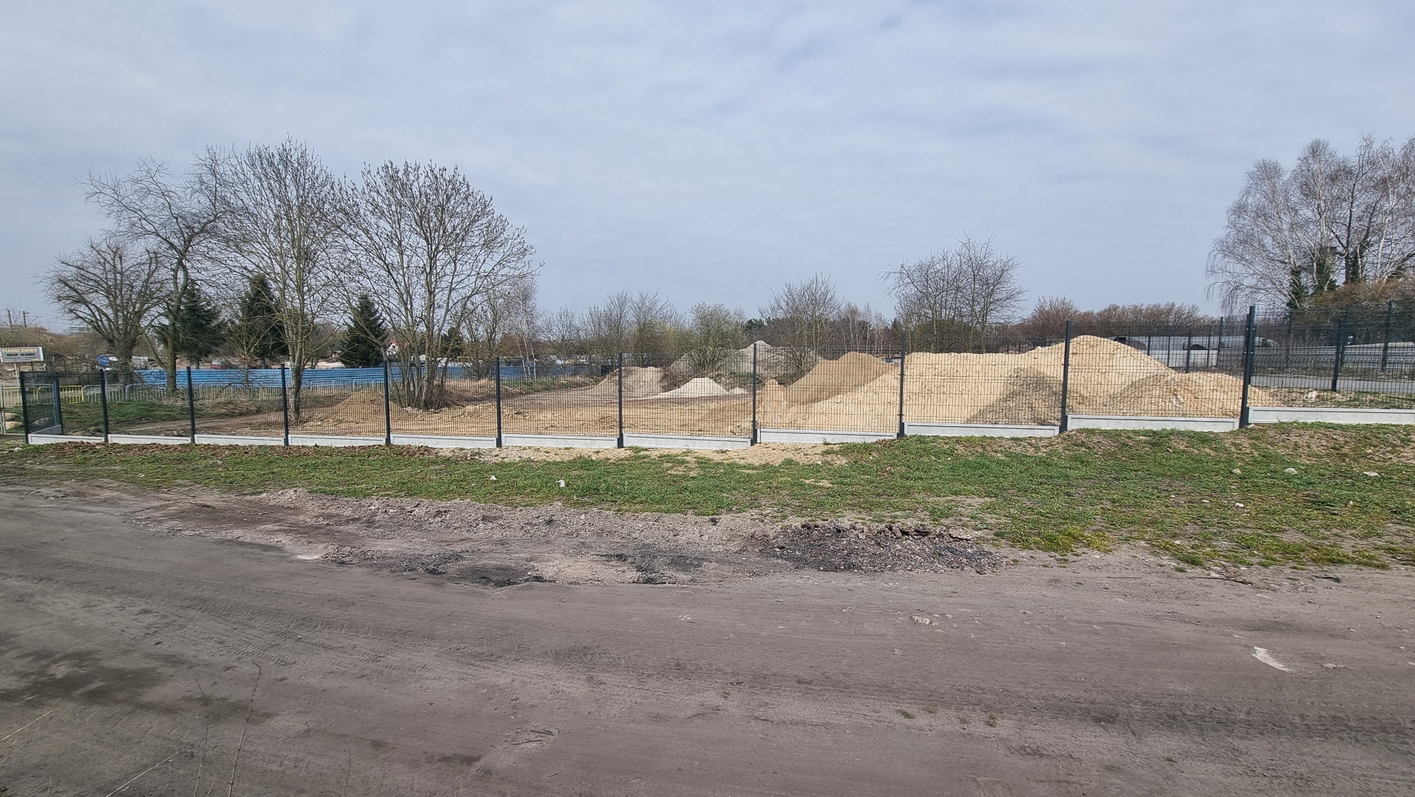 Teren budowy z pryzmami piasku za metalowym ogrodzeniem panelowym na betonowej podmurówce. W tle drzewa bez liści i błękitne bariery. Pochmurne niebo.