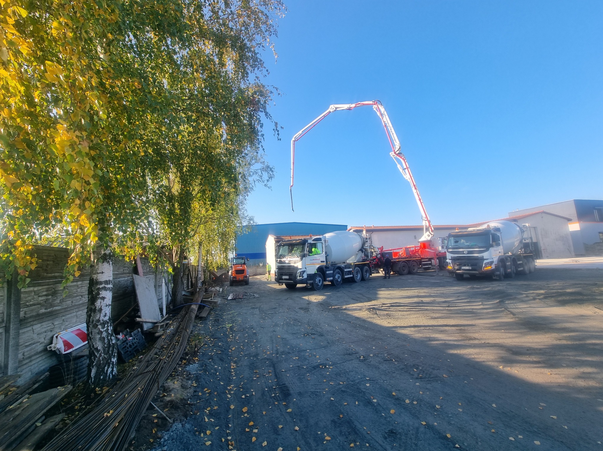 Betonomieszarki z pompą na placu budowy, widoczne zbrojenia i jesienne drzewo. Słoneczny dzień, błękitne niebo. Charakterystyczny wysięgnik pompy do betonu.