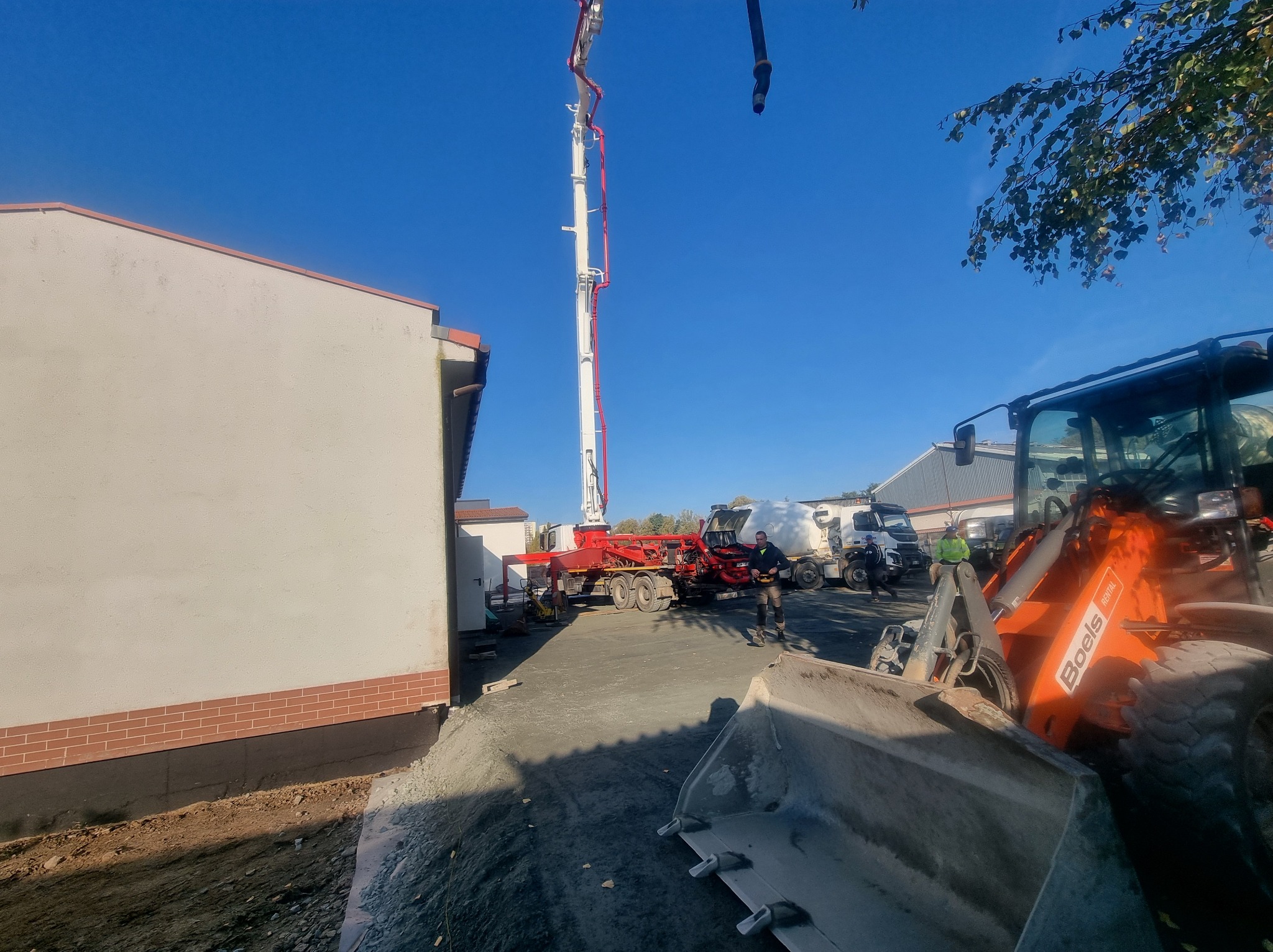 Betoniarka pompuje beton na budowę domu jednorodzinnego. Widoczny operator, koparka i fragment budynku z ceglaną podmurówką. Błękitne niebo w tle.