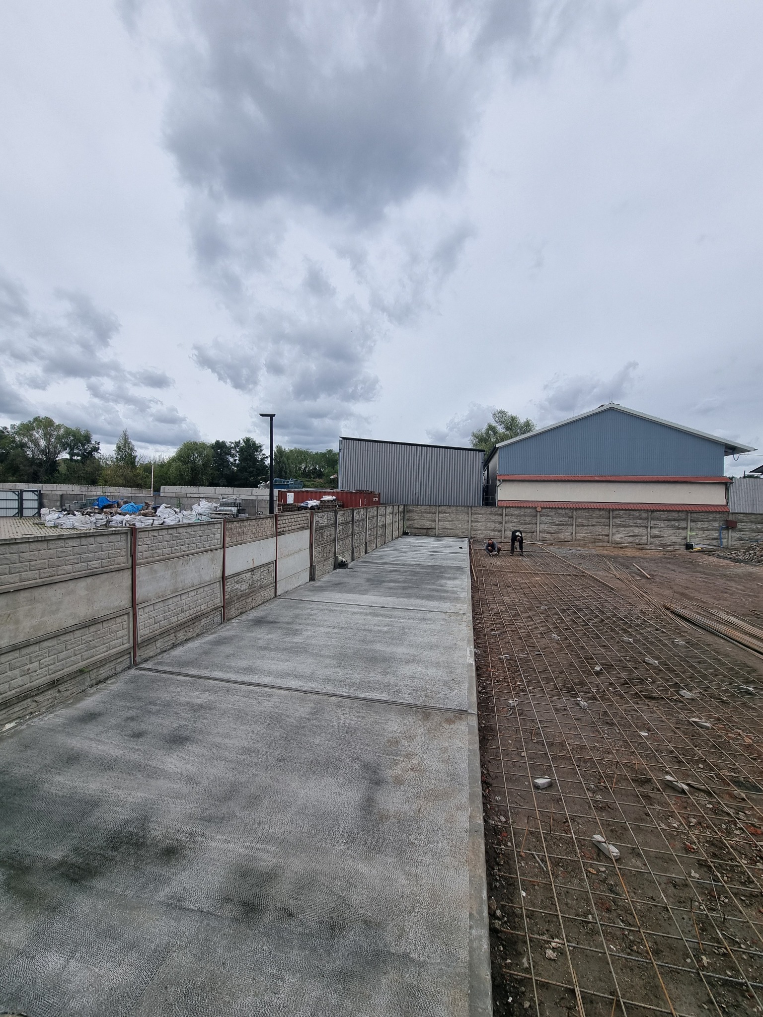 Betonowa wylewka na placu budowy, widoczne zbrojenia i pracownicy w tle. Szare niebo i budynki w tle. Betonowa ściana oddzielająca teren.