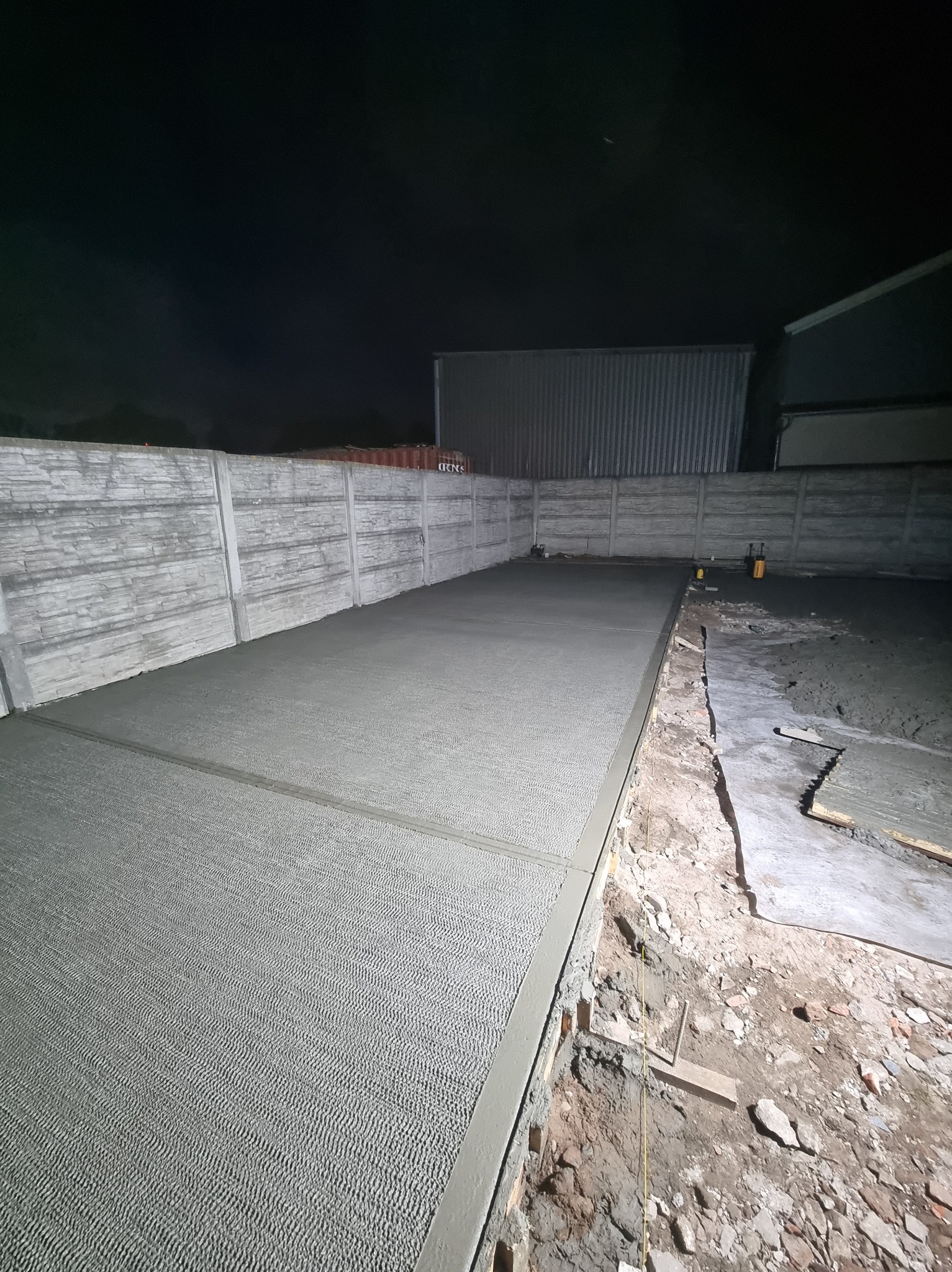 Świeżo wylana, szara wylewka betonowa na zewnątrz, otoczona betonowym ogrodzeniem. Część wylewki jeszcze nieukończona, widoczne narzędzia i elementy szalunku.