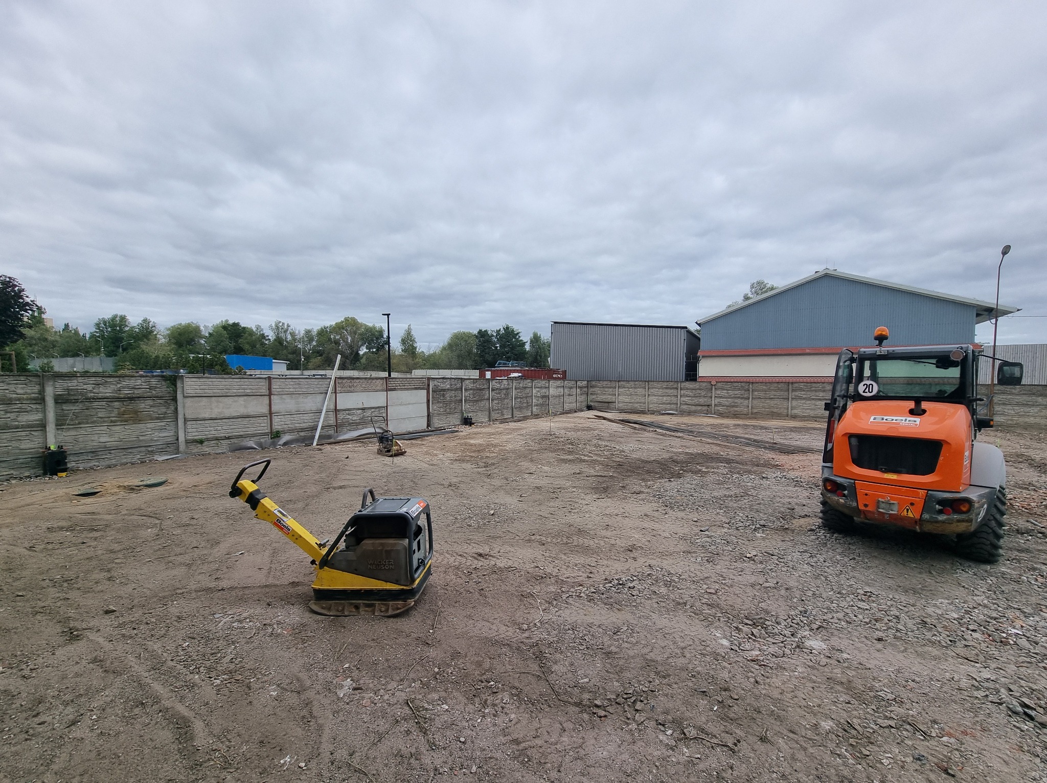Teren budowy z ubijarką i minikoparką na ubitej ziemi, ogrodzony betonowym płotem. W tle budynki i pochmurne niebo. Widok na przygotowany plac pod budowę.
