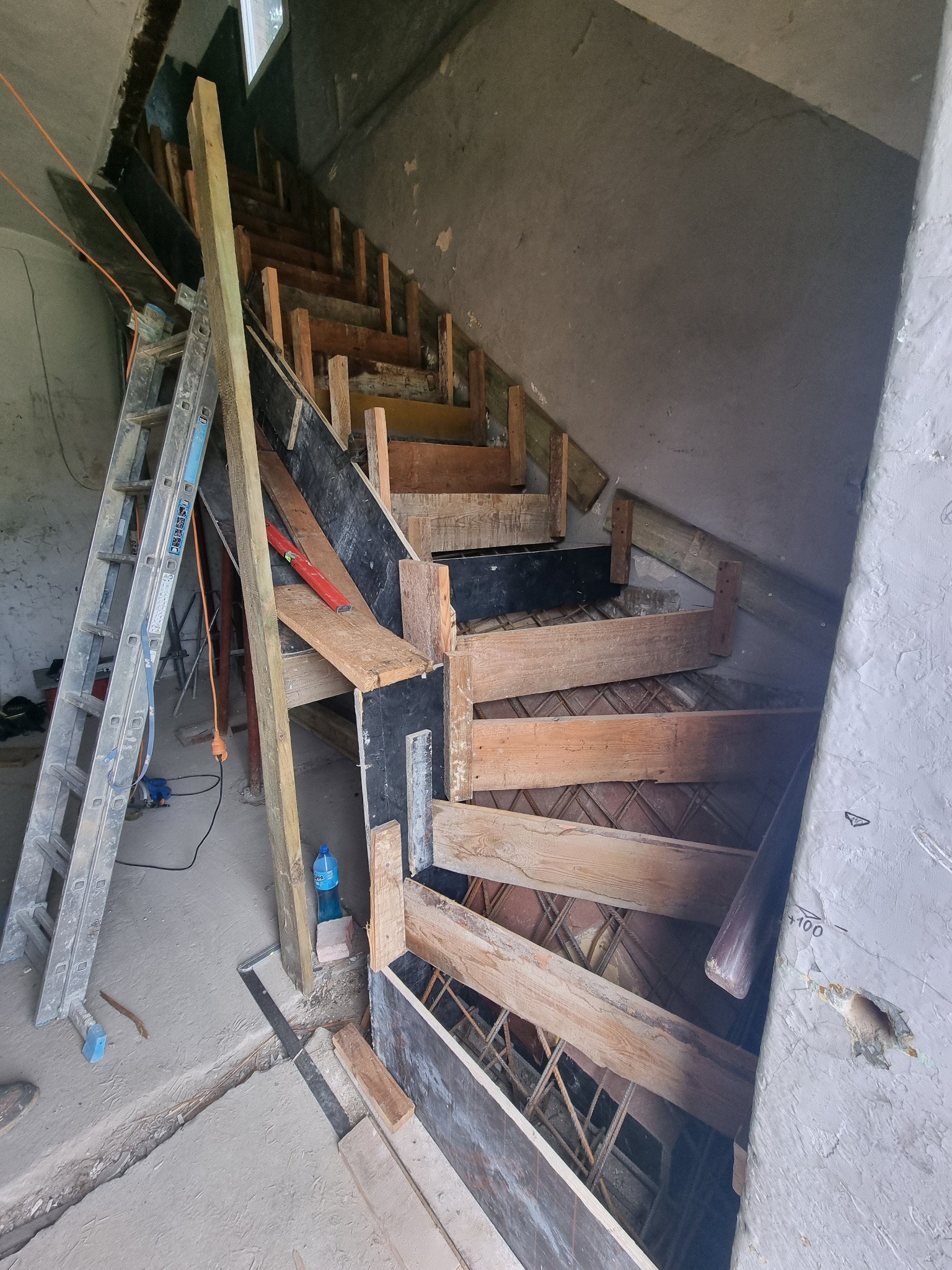 Wnętrze w trakcie budowy schodów spiralnych z drewnianym szalunkiem i zbrojeniem, widok z dołu, drabina oparta o ścianę, surowy stan wykończenia.