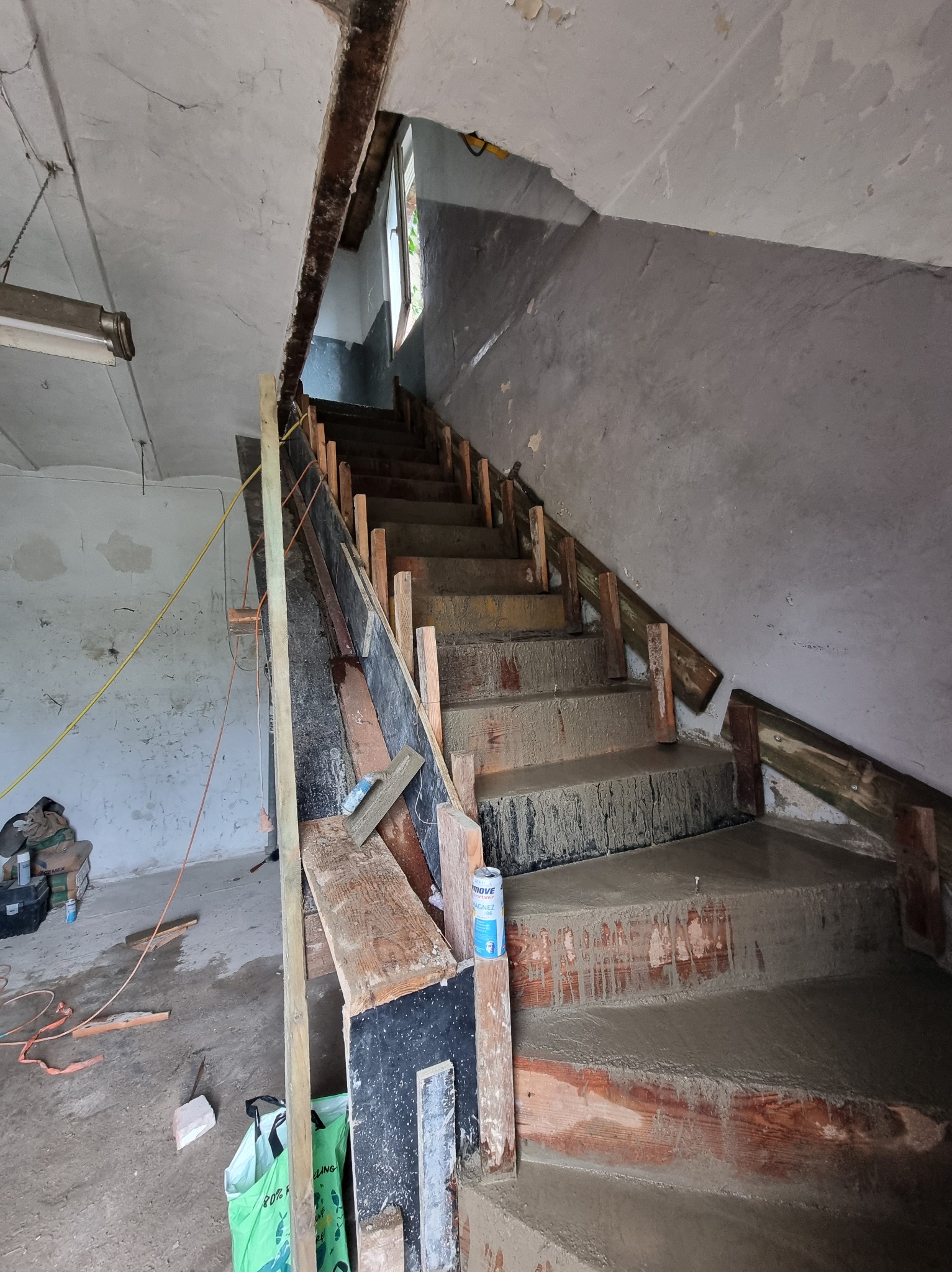 Wnętrze budynku w trakcie remontu: betonowe schody z drewnianym szalunkiem, widoczne narzędzia i materiały budowlane. Surowy stan wykończenia ścian i sufitu.