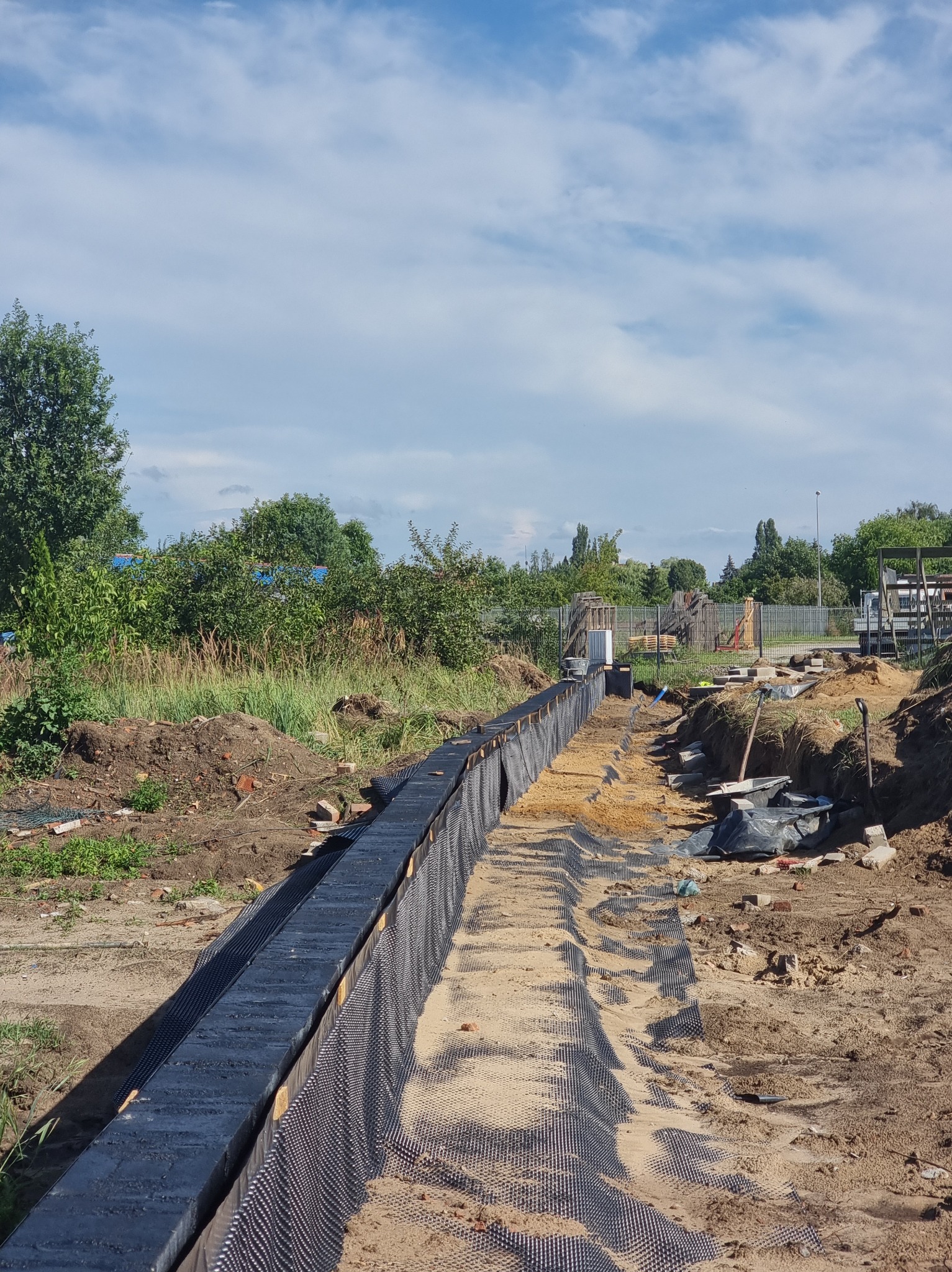 Przygotowanie wykopu pod fundament z izolacją pionową. Widoczne szalunki i folia kubełkowa. Prace ziemne w toku, teren budowy z roślinnością w tle.