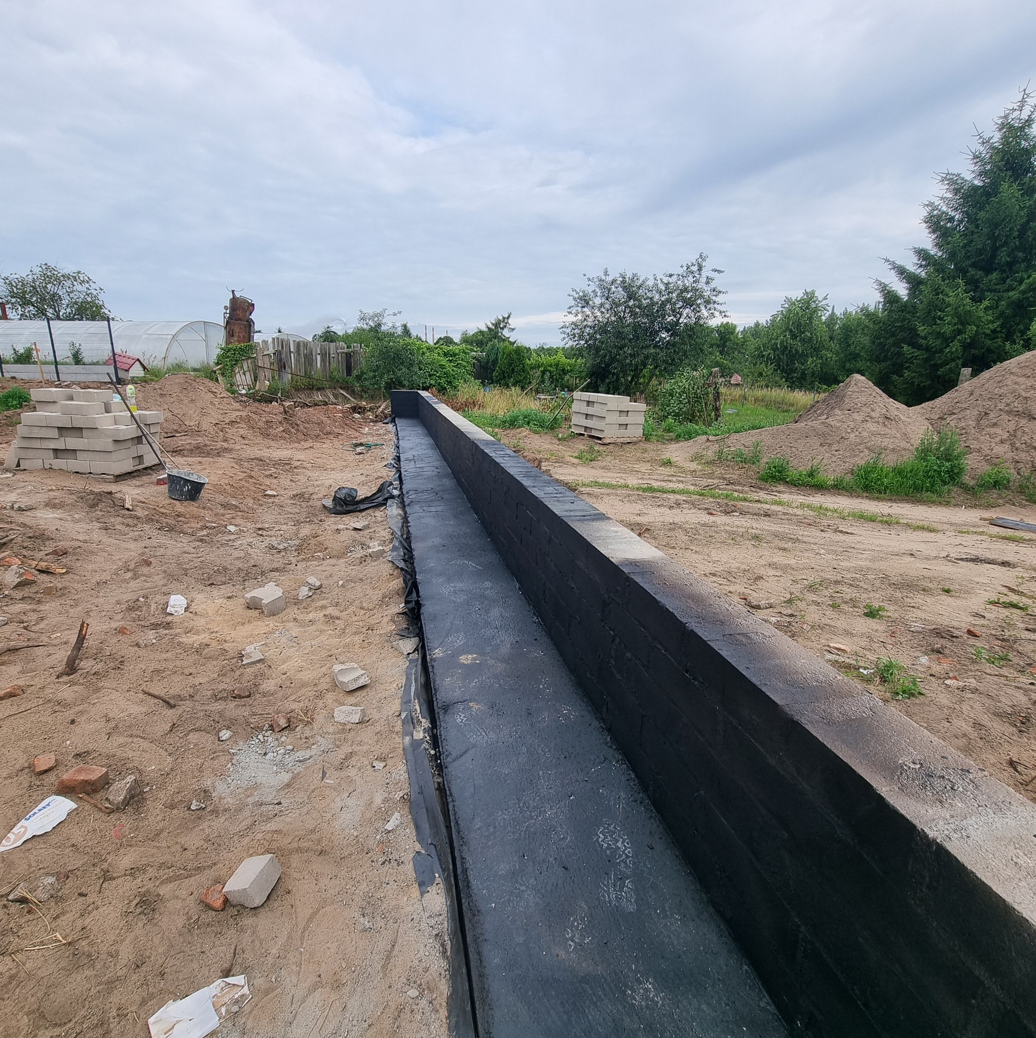 Zabezpieczone fundamenty pomalowane na czarno, widoczne bloczki betonowe i narzędzia na placu budowy. Tło z zielenią i pochmurnym niebem.