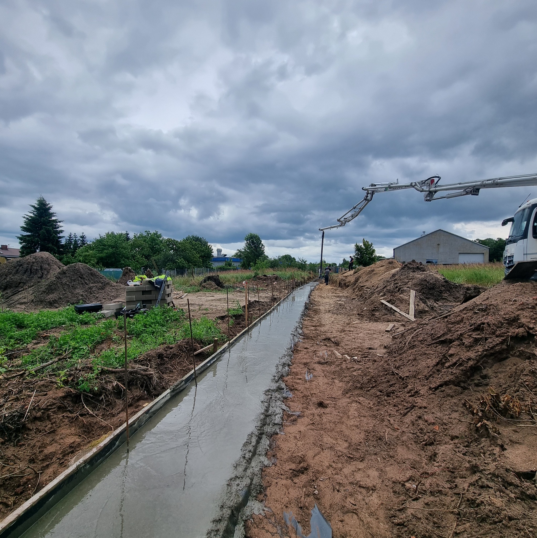 Wylewka fundamentowa w szalunku, beton rozprowadzany pompą. Widoczne zbrojenie i przygotowany teren budowy pod pochmurnym niebem.