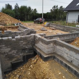 Len-Bud - Budowa fundamentów domu jednorodzinnego z bloczków betonowych, widoczne ściany fundamentowe, izolacja pionowa, wykop, nasyp ziemi i zaparkowany samochód w tle.