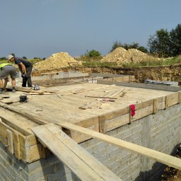 Len-Bud - Dwóch pracowników budowlanych pochylonych nad szalunkiem fundamentu z cegieł, widoczne drewniane deski, wystające pręty zbrojeniowe, w tle hałda piasku i wykop ziemny.