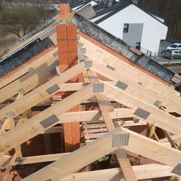 Len-Bud - Widok z góry na konstrukcję dachu z drewnianych belek i komin z czerwonej cegły w trakcie budowy.