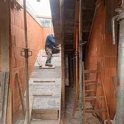 Len-Bud - Budowa domu: murarz pracuje na drewnianym rusztowaniu wewnątrz budynku, widoczne ściany z czerwonej cegły, podpory i drewniana drabina.