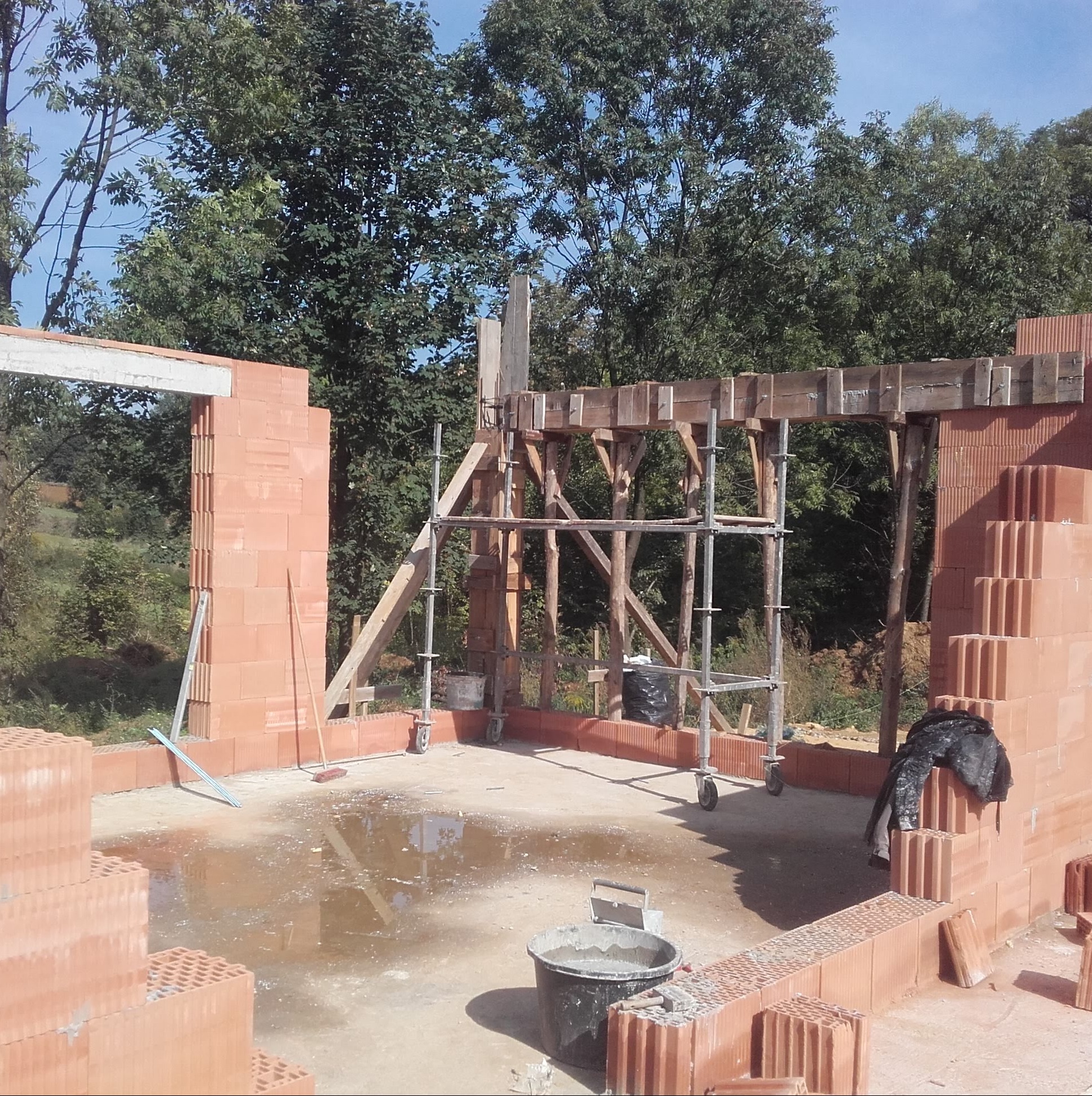 Wznoszenie ścian domu z pustaków ceramicznych, widoczne rusztowanie, wiadro z zaprawą i kałuże na podłożu.