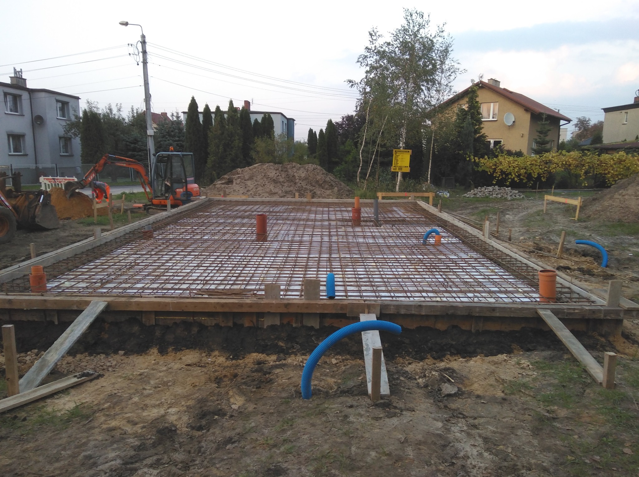 Zbrojenie fundamentu domu jednorodzinnego, widoczne szalunki i rury instalacyjne, w tle koparka i zabudowa mieszkalna.