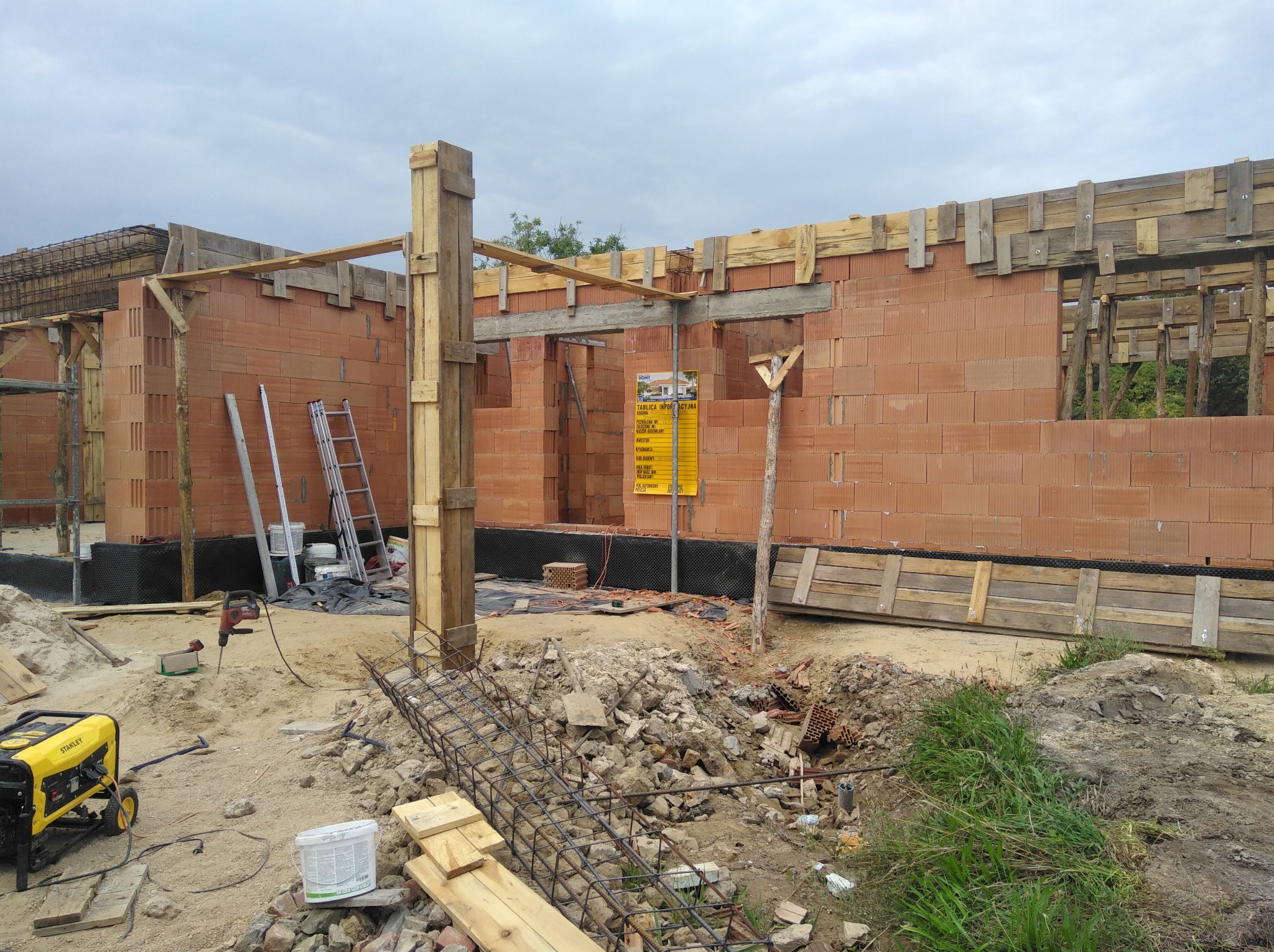 Budowa domu z czerwonej cegły porotherm, widoczne drewniane szalunki, zbrojenie, generator prądu Stanley i drabiny na placu budowy