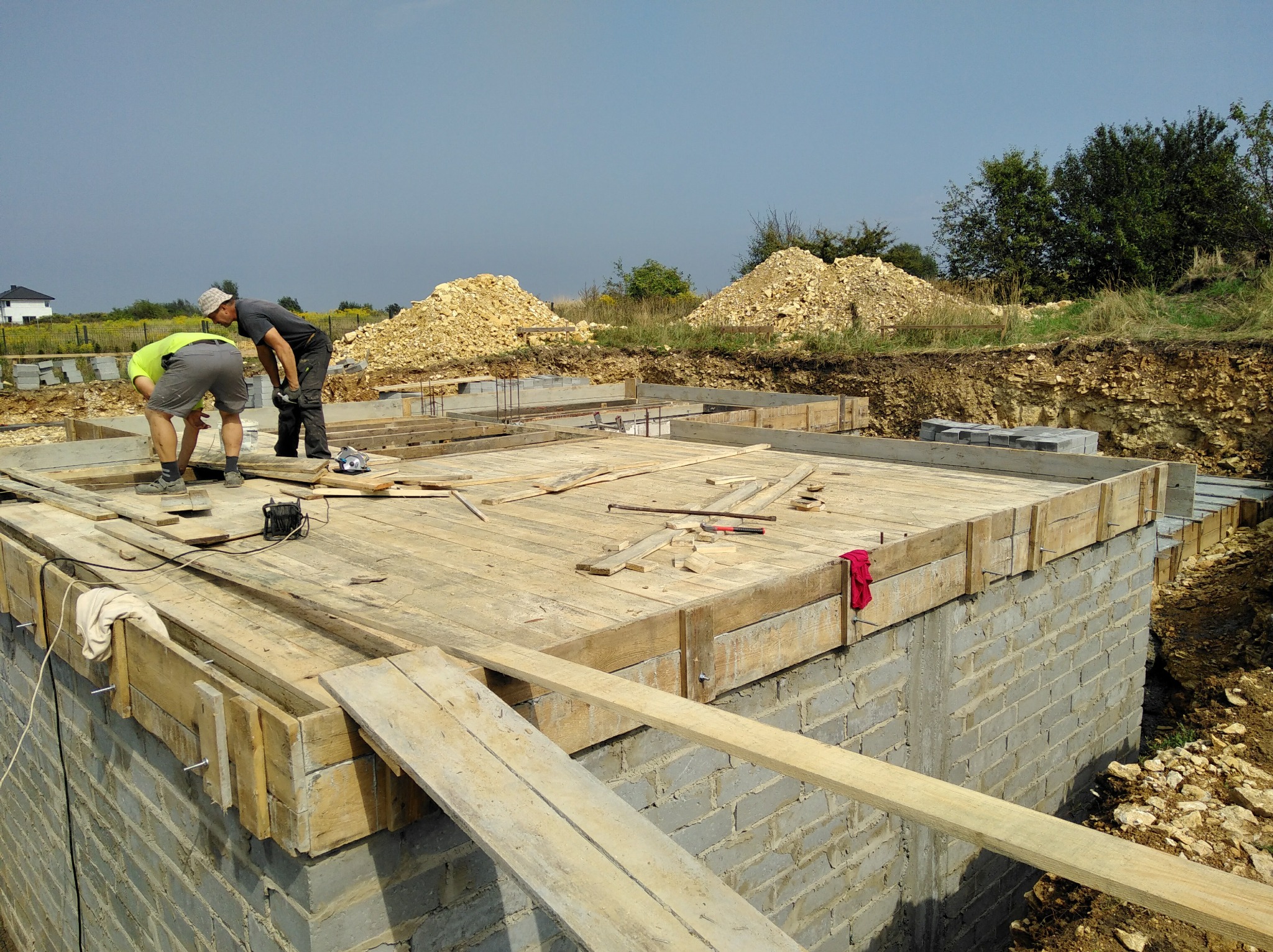 Dwóch pracowników budowlanych pochylonych nad szalunkiem fundamentu z cegieł, widoczne drewniane deski, wystające pręty zbrojeniowe, w tle hałda piasku i wykop ziemny.