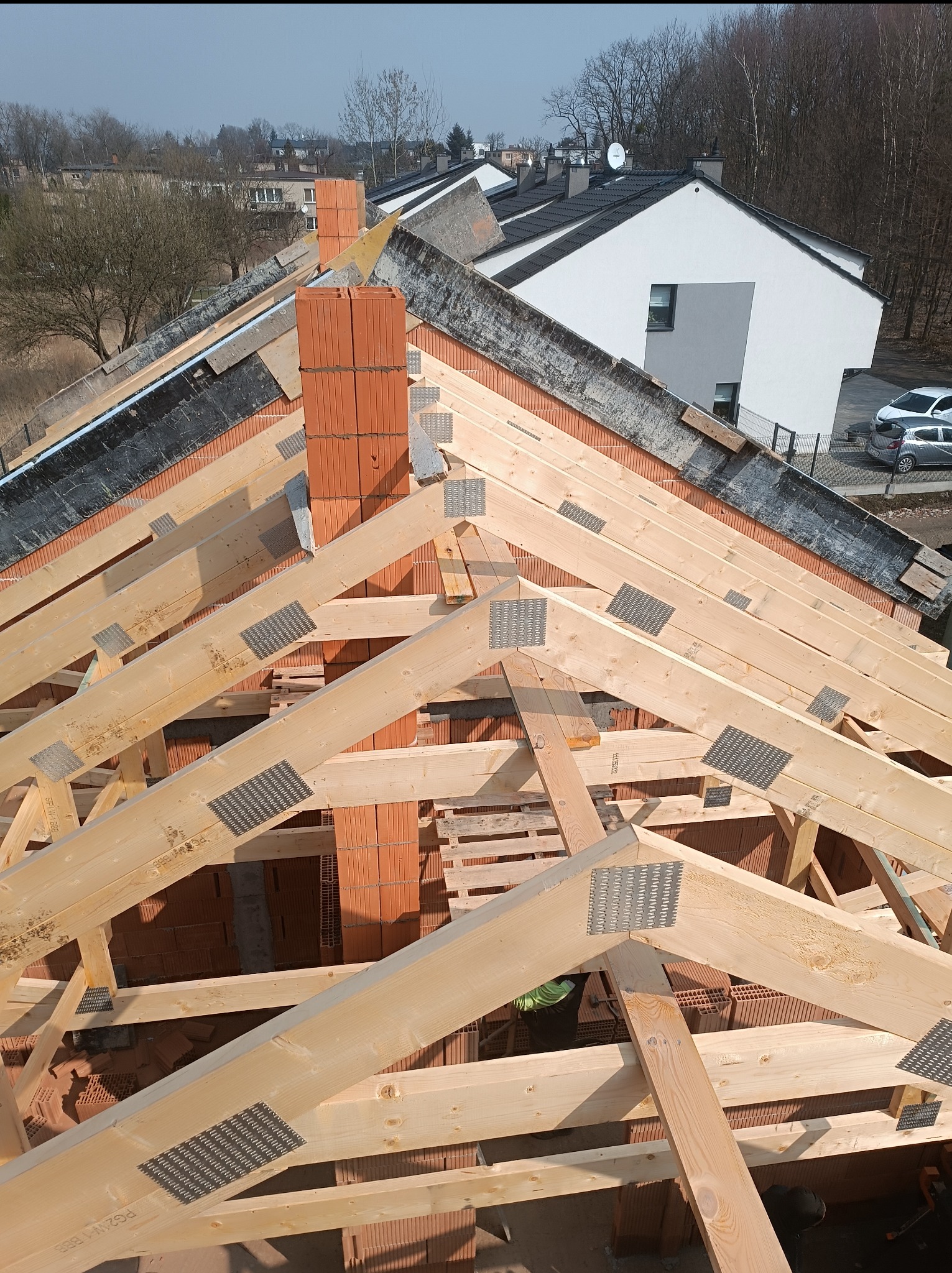 Widok z góry na konstrukcję dachu z drewnianych belek i komin z czerwonej cegły w trakcie budowy.